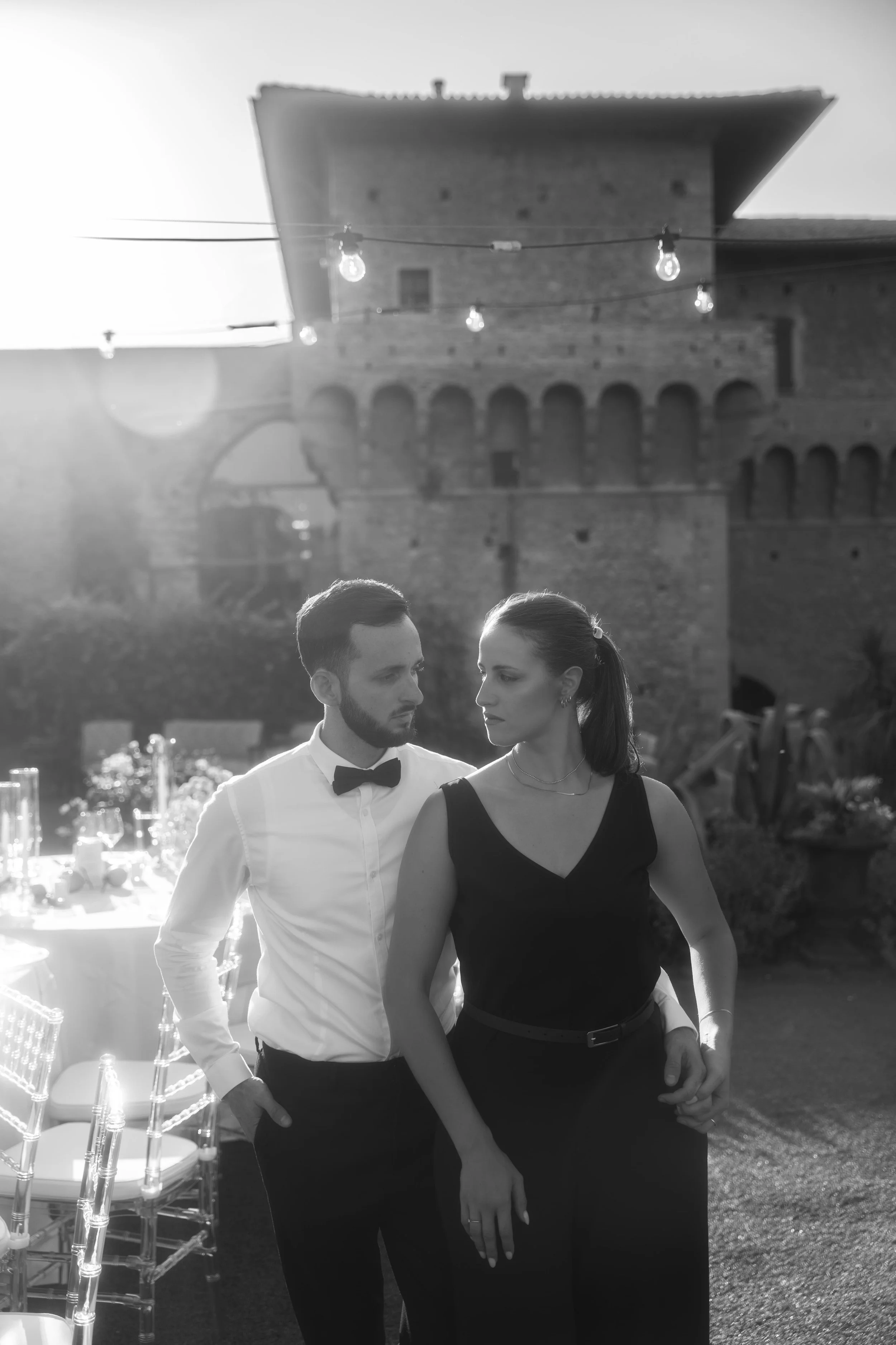 Una coppia elegante, un uomo e una donna, si ritrovano in un evento all'aperto al tramonto, con decorazioni di luci e tavoli apparecchiati.