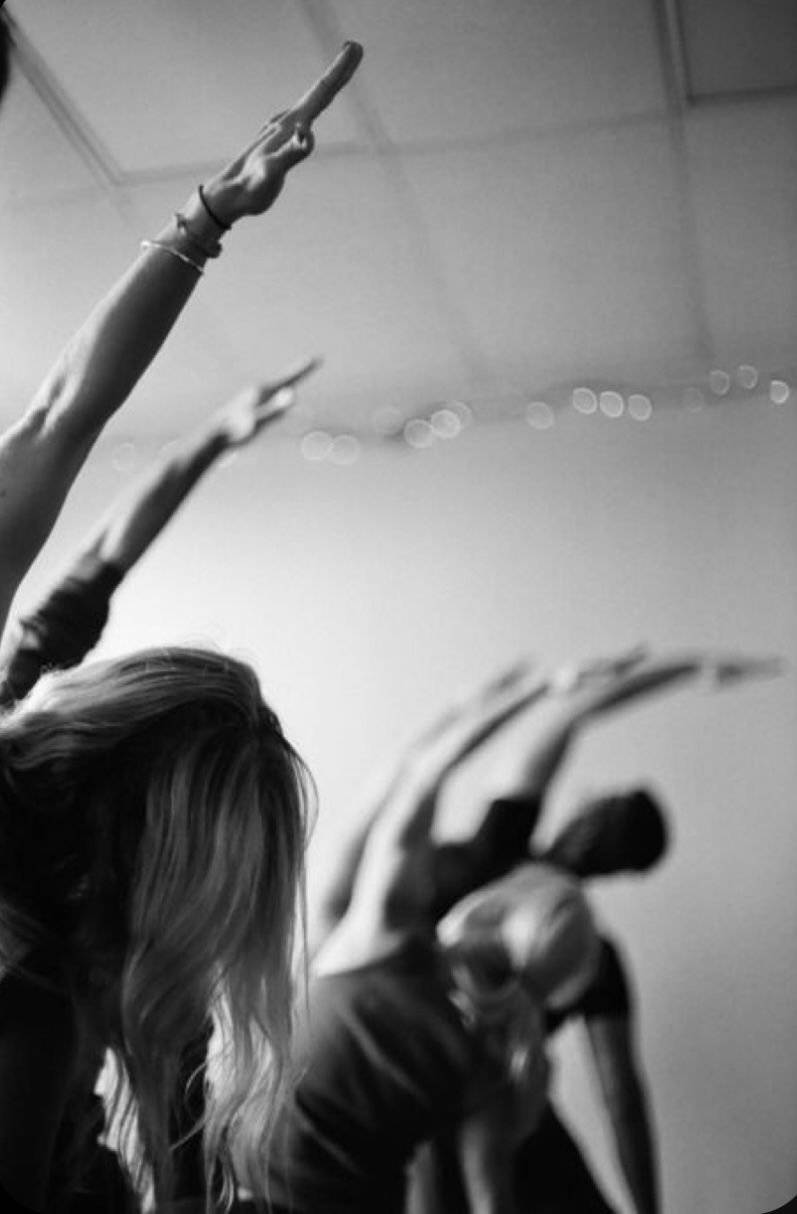 People participating in a yoga or stretching class, with their arms raised and bent at the waist, in a studio with string lights on the ceiling.