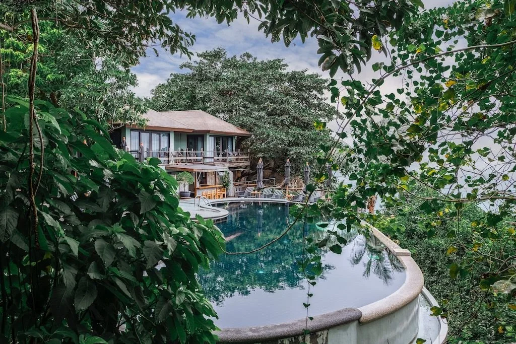 A tropical overwater bungalow near a swimming pool, surrounded by dense green jungle foliage, with trees and a cloudy sky in the background.