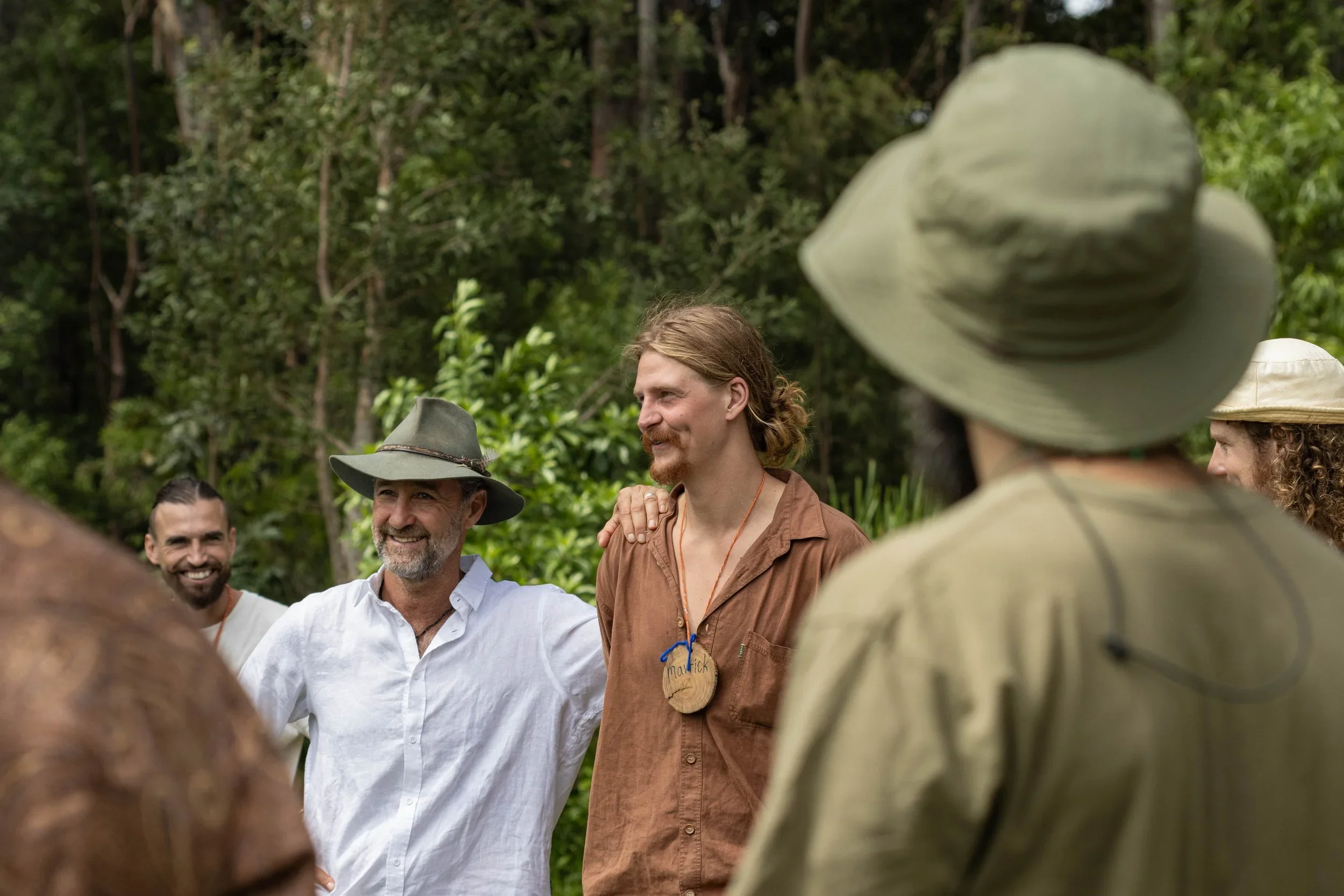 Group of people outdoors, standing in a circle, smiling, some wearing hats, surrounded by green trees.