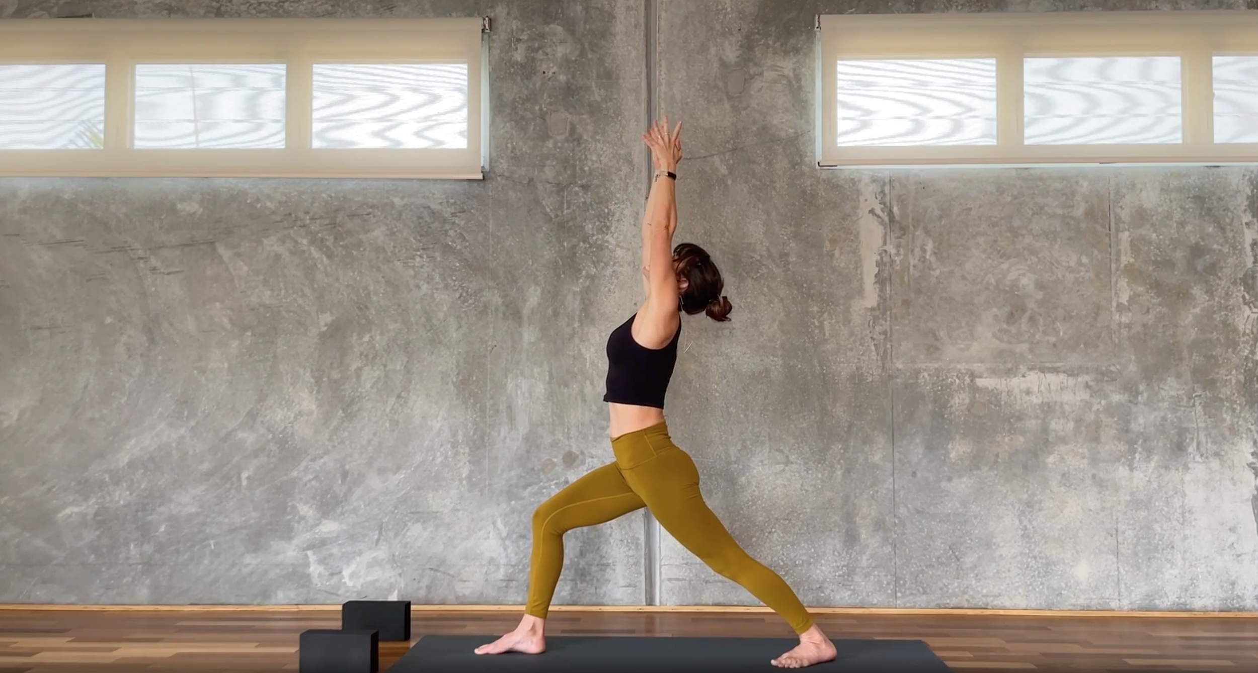 A woman practicing yoga in a studio with a concrete wall background and windows at the top. She is in a high lunge pose with arms raised overhead, wearing a black sleeveless top and olive green leggings.