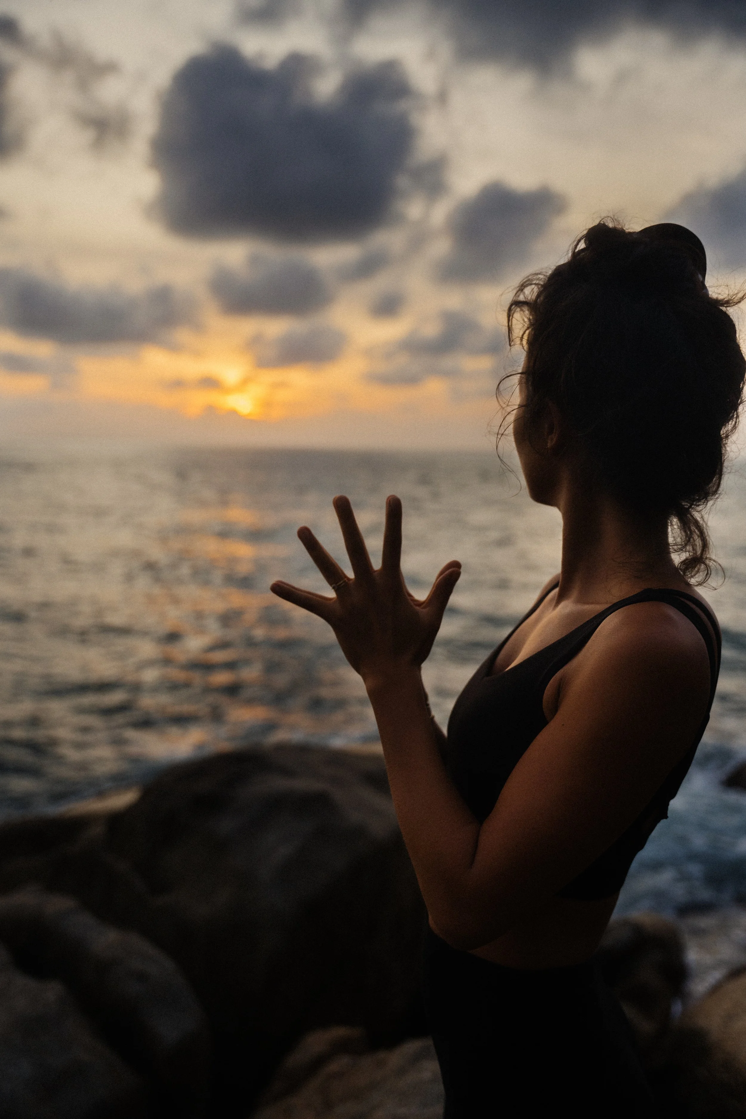 A woman with her hair tied up observing a sunset over the ocean, standing next to rocks.