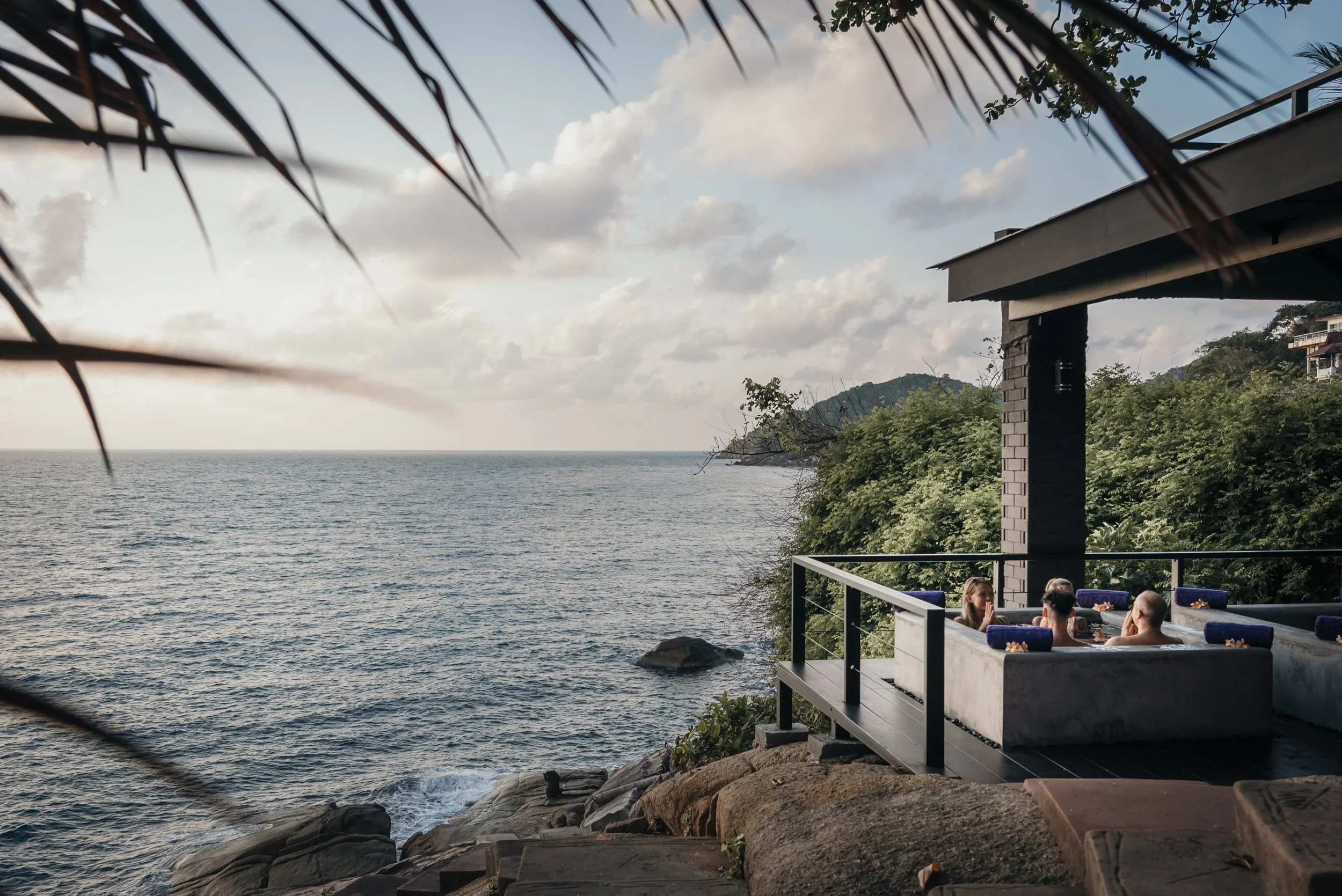 People relaxing in a hot tub on a rocky coastline with the ocean and cloudy sky in the background.