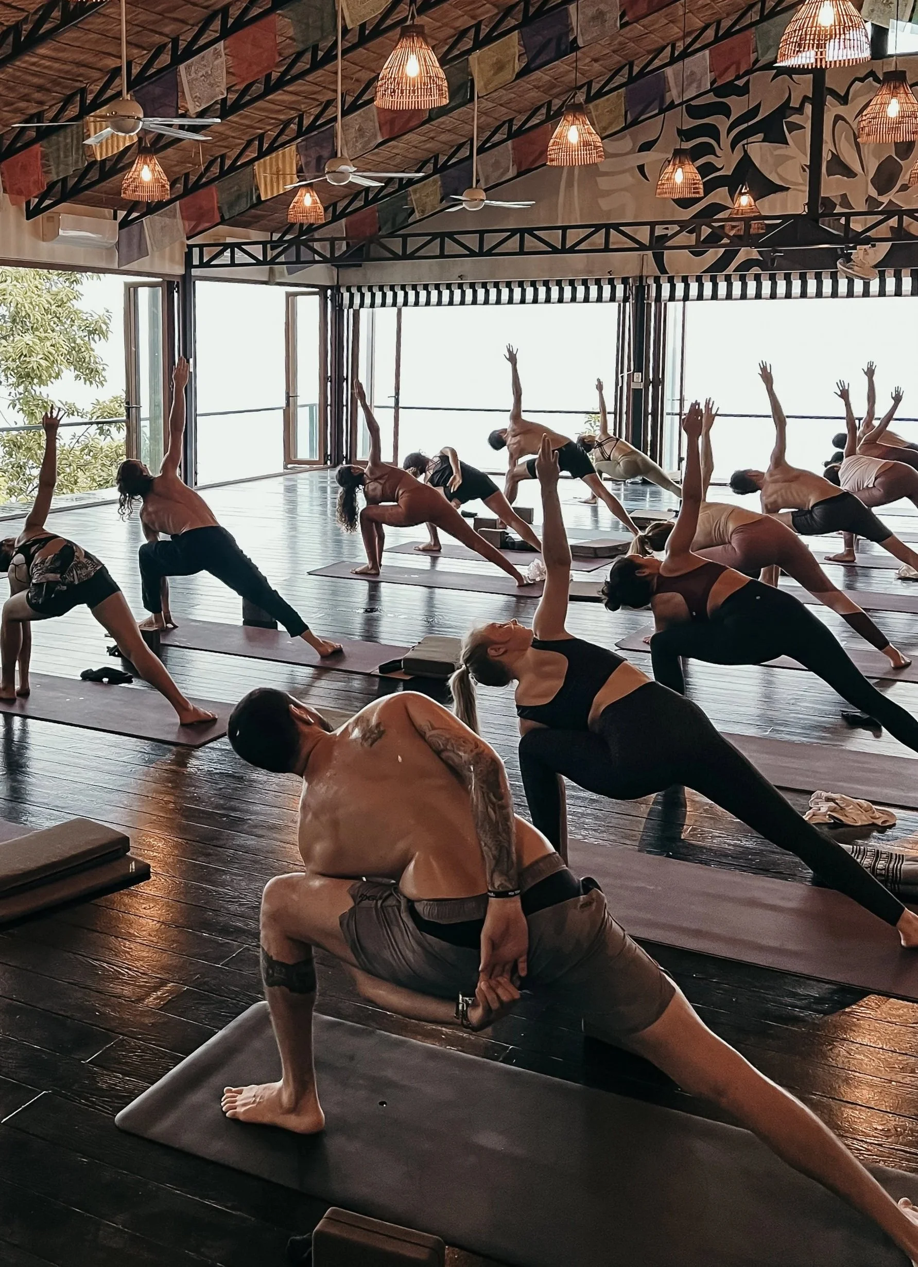 People participating in a yoga class in a bright studio with large windows, wooden floors, and hanging lights.