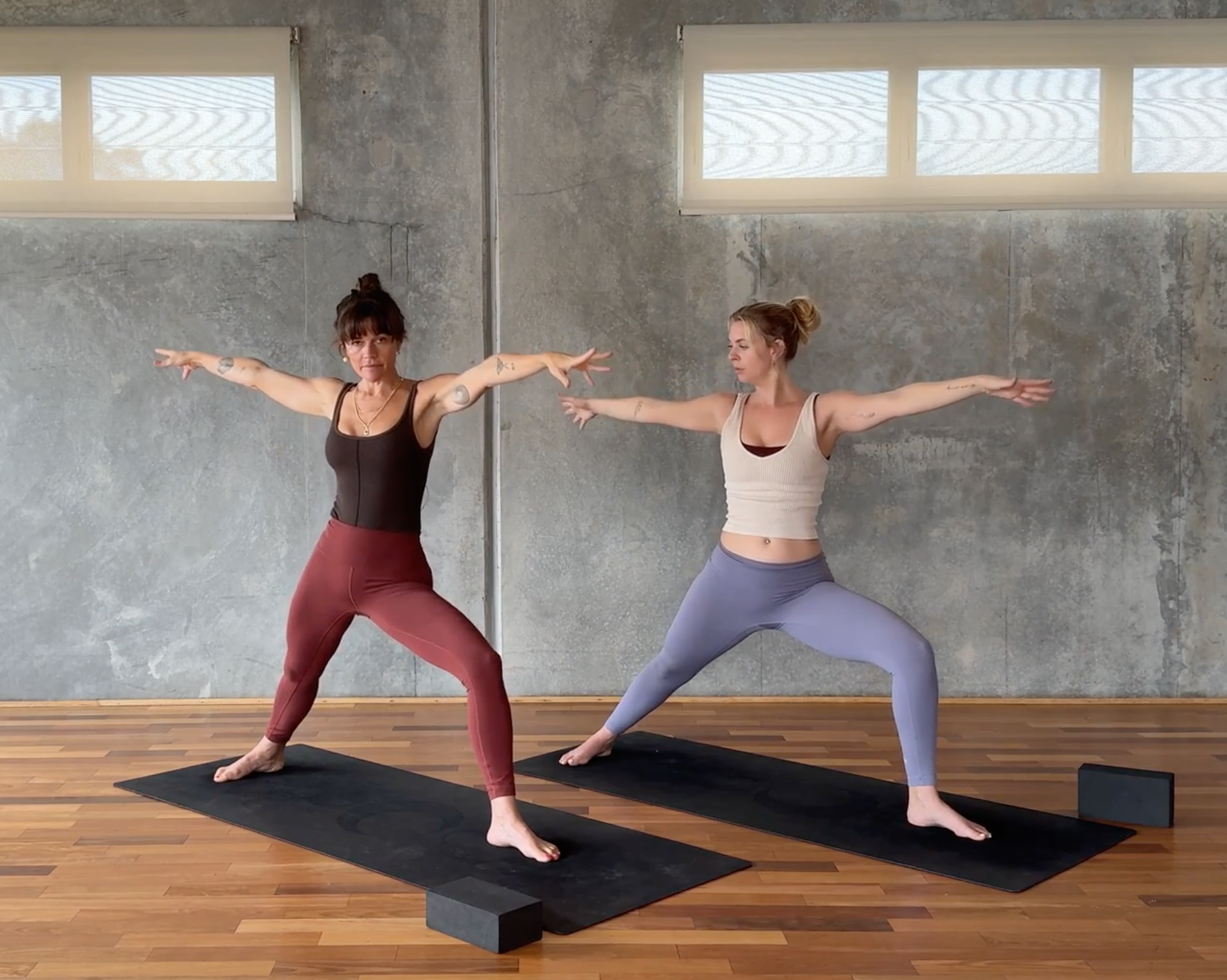 Two women practicing yoga in a studio, standing on black mats with yoga blocks, arms extended sideways, balancing and stretching.