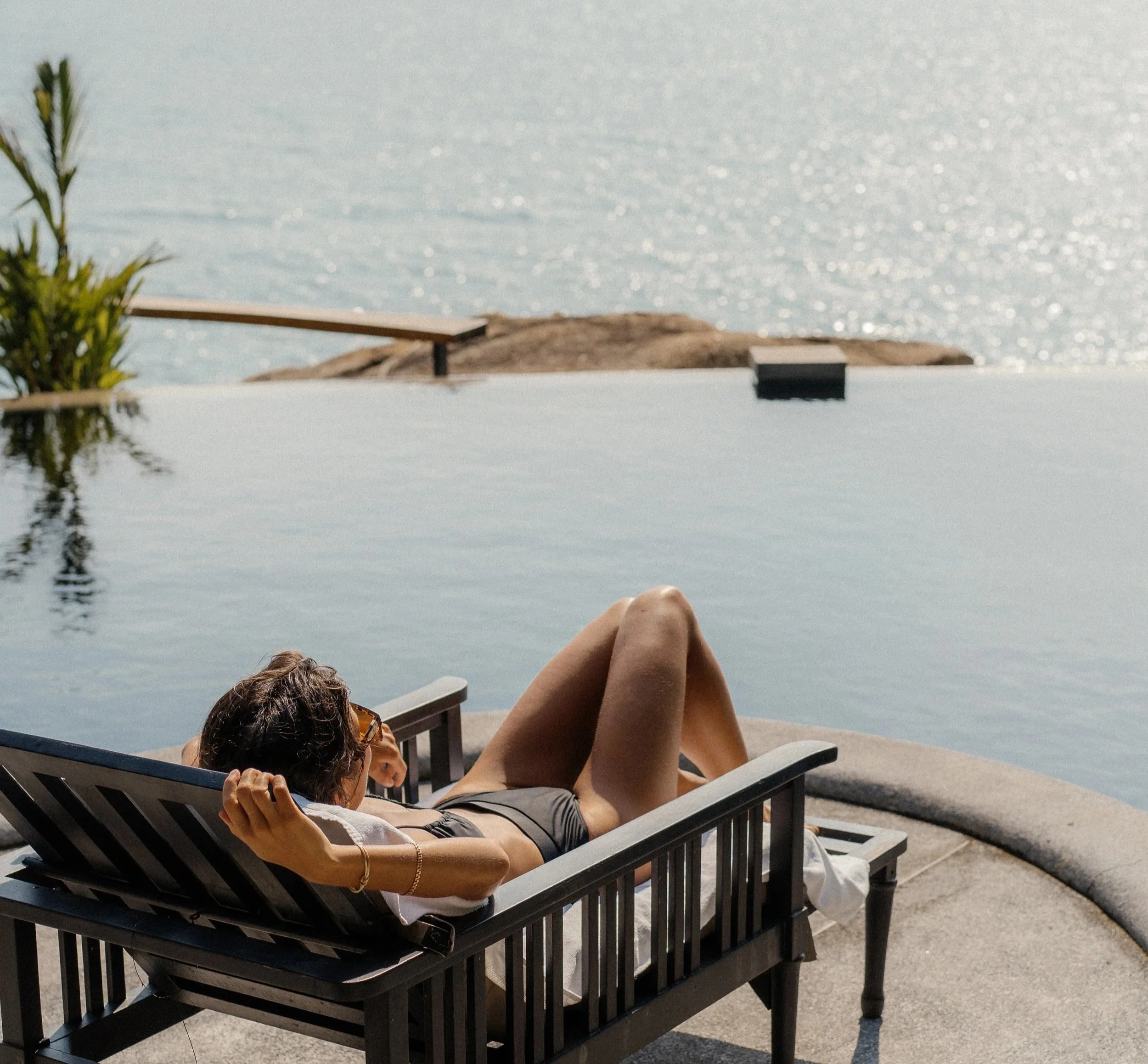 A woman relaxing on a lounge chair by a swimming pool overlooking the water, wearing sunglasses and a black swimsuit.