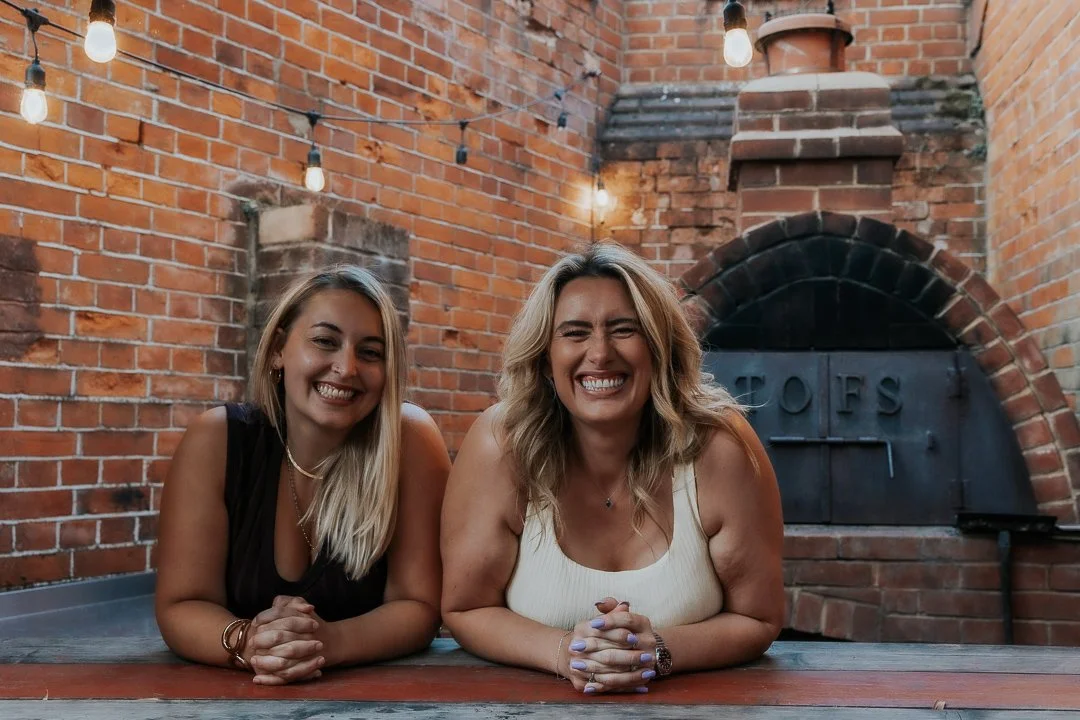 Two women smiling and leaning on an outdoor table with his hands clasped, in front of a brick wall and an oven