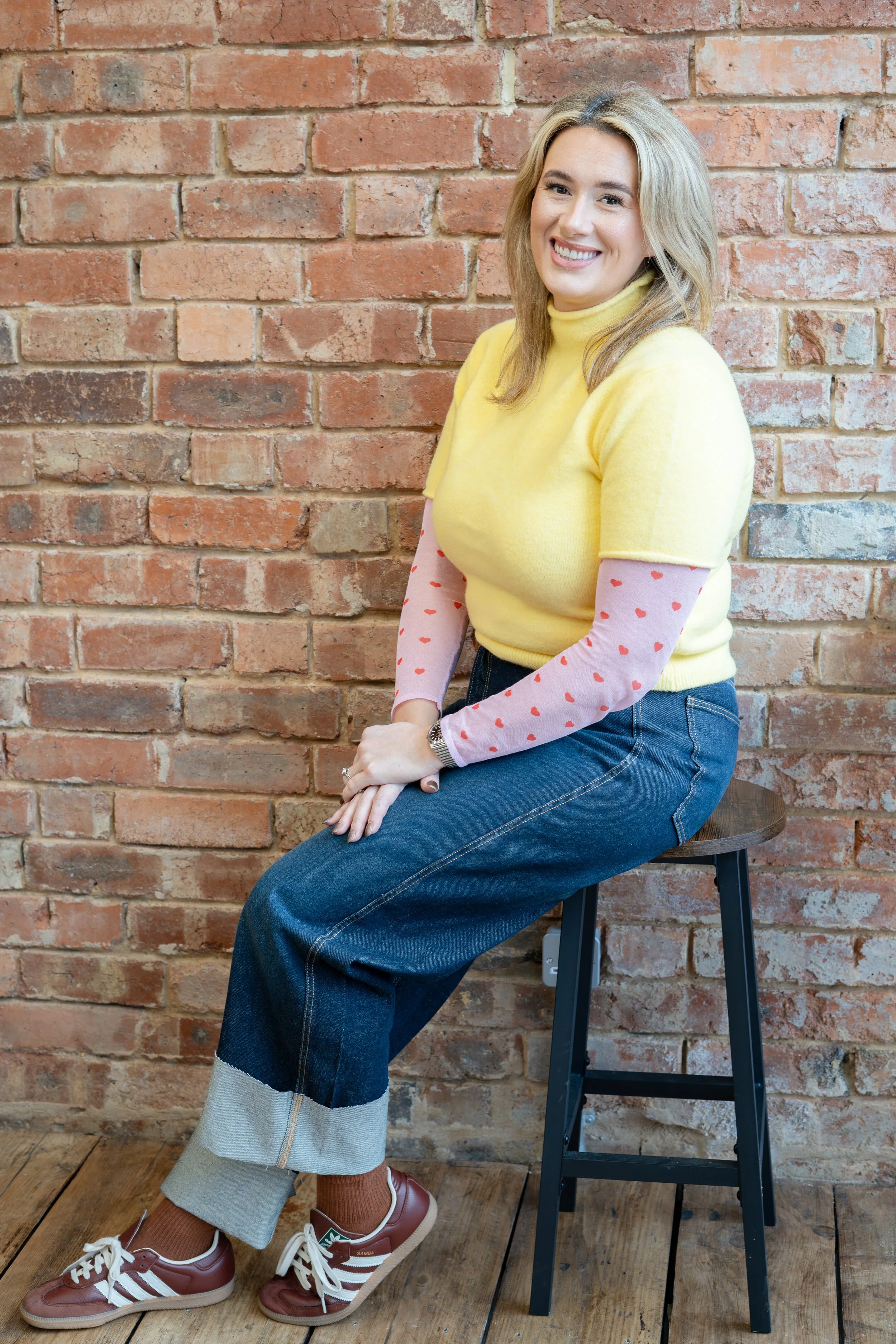 A smiling woman with blonde hair sitting on a black stool against a red brick wall, wearing a yellow sweater with pink sleeves that have red hearts, blue jeans, brown sneakers with white laces, and brown socks.