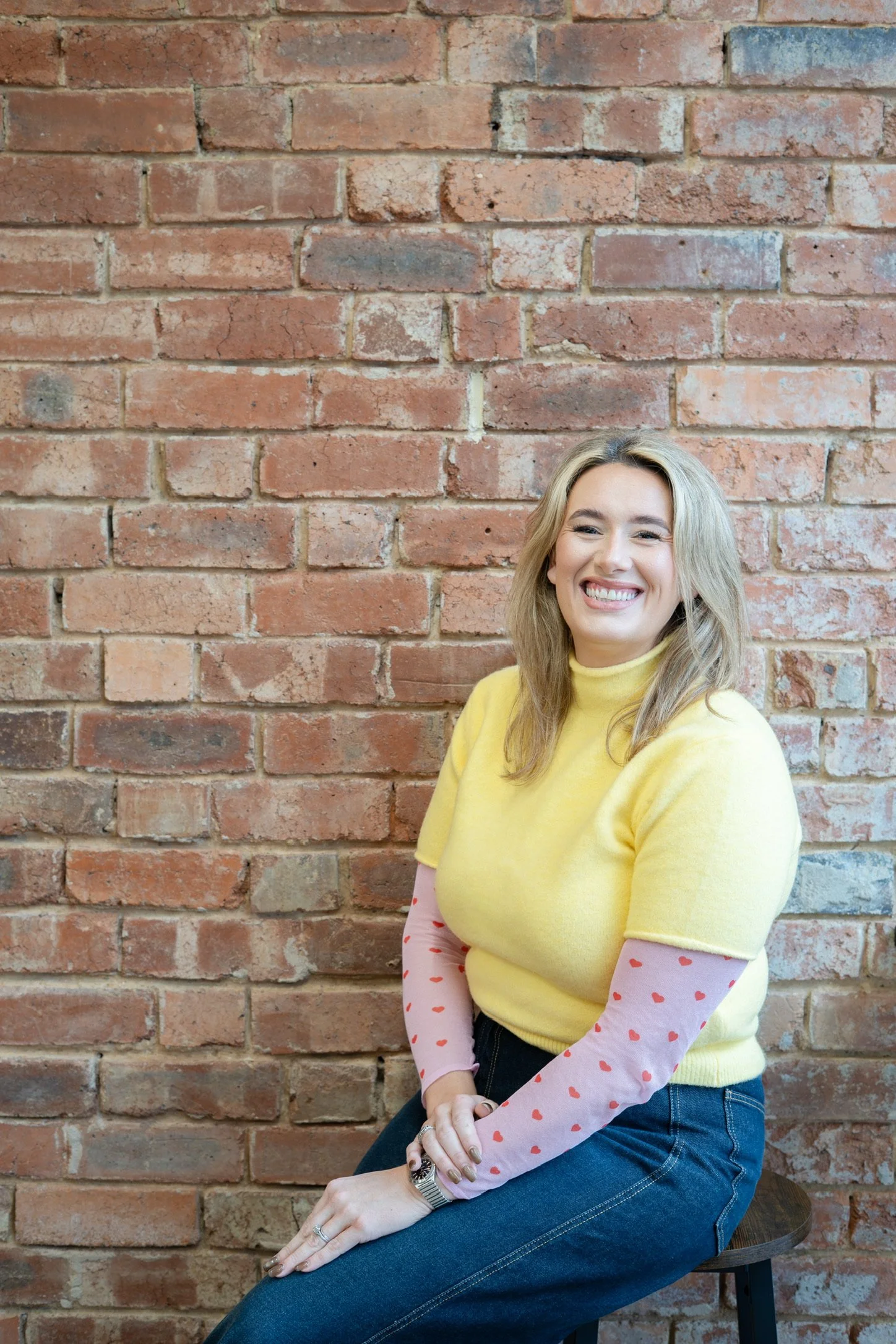 A woman with blonde hair and a smile, wearing a yellow sweater over pink long sleeves with red hearts, sitting on a stool against a brick wall.