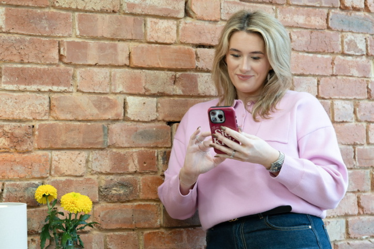 A woman with blonde hair wearing a pink sweatshirt and dark jeans looks at her phone while standing against a red brick wall.