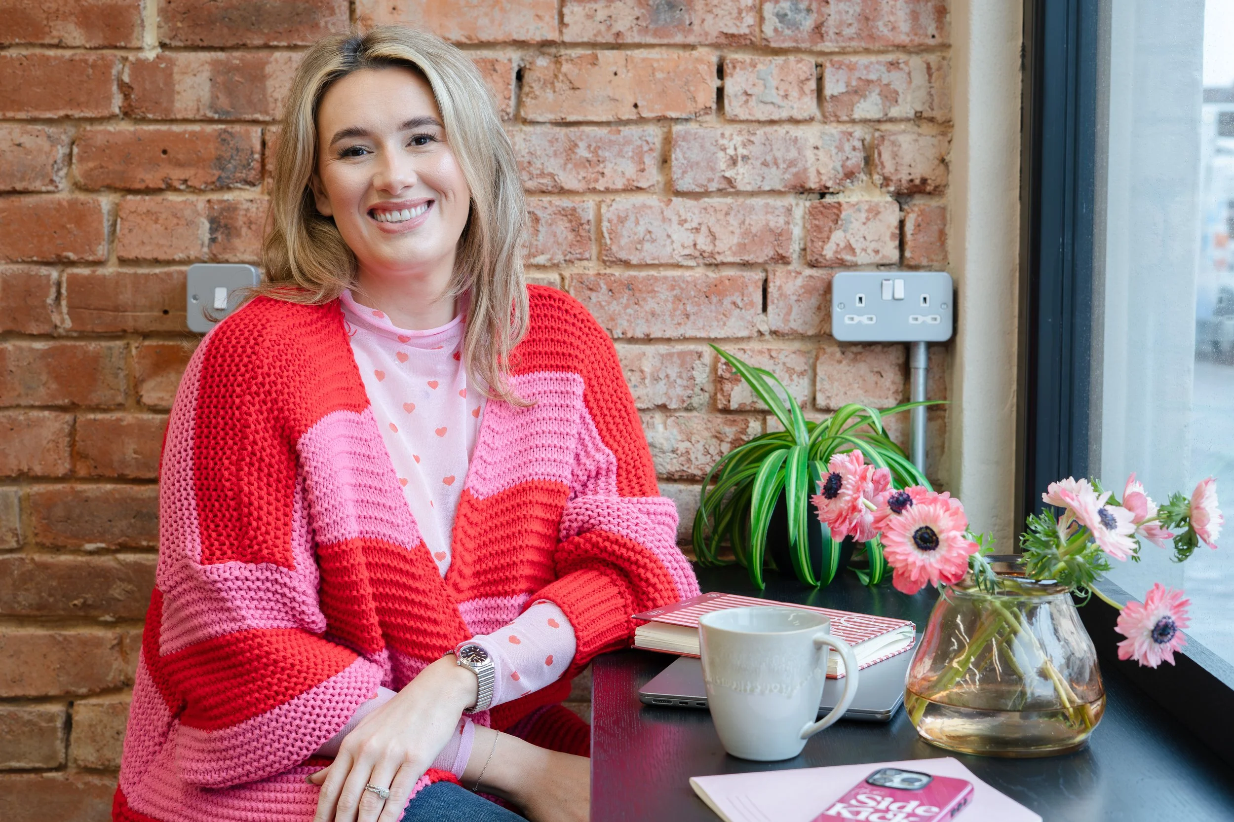 A smiling woman with blonde hair wearing a pink and red knitted sweater sitting at a table with a flower arrangement, a white mug, a pink notebook, and a phone in front of her, against a brick wall and window background.