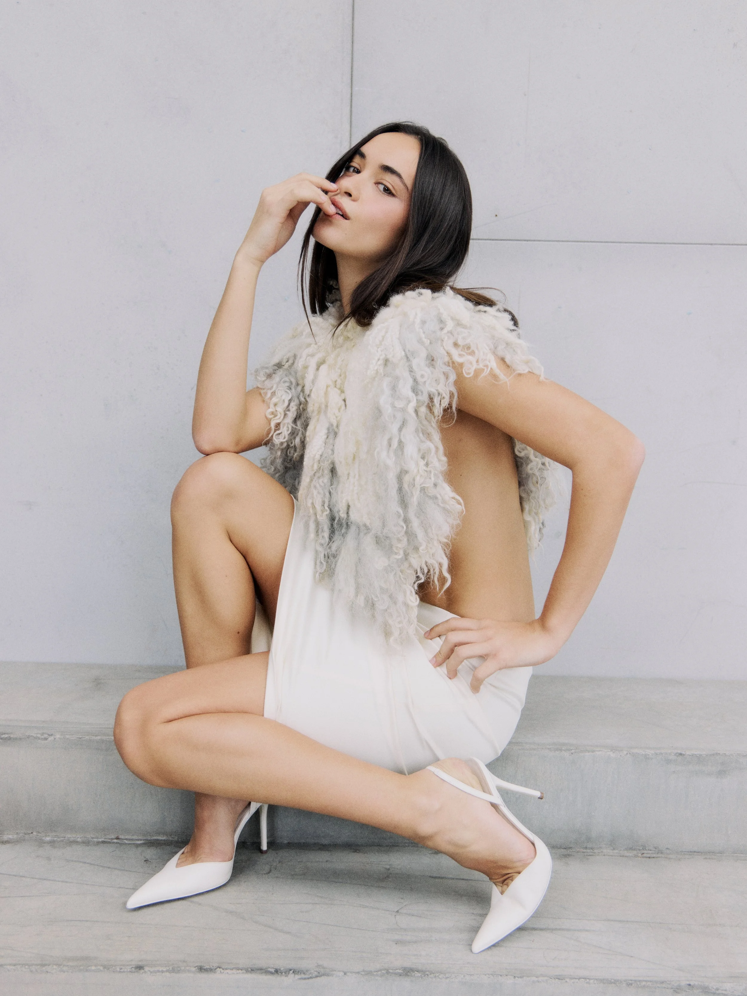 A woman with dark hair and light skin, wearing a fluffy cream-colored top and a white skirt, is sitting on a step against a plain wall. She has her hand near her face and is wearing white high heels. Her pose is confident and relaxed.