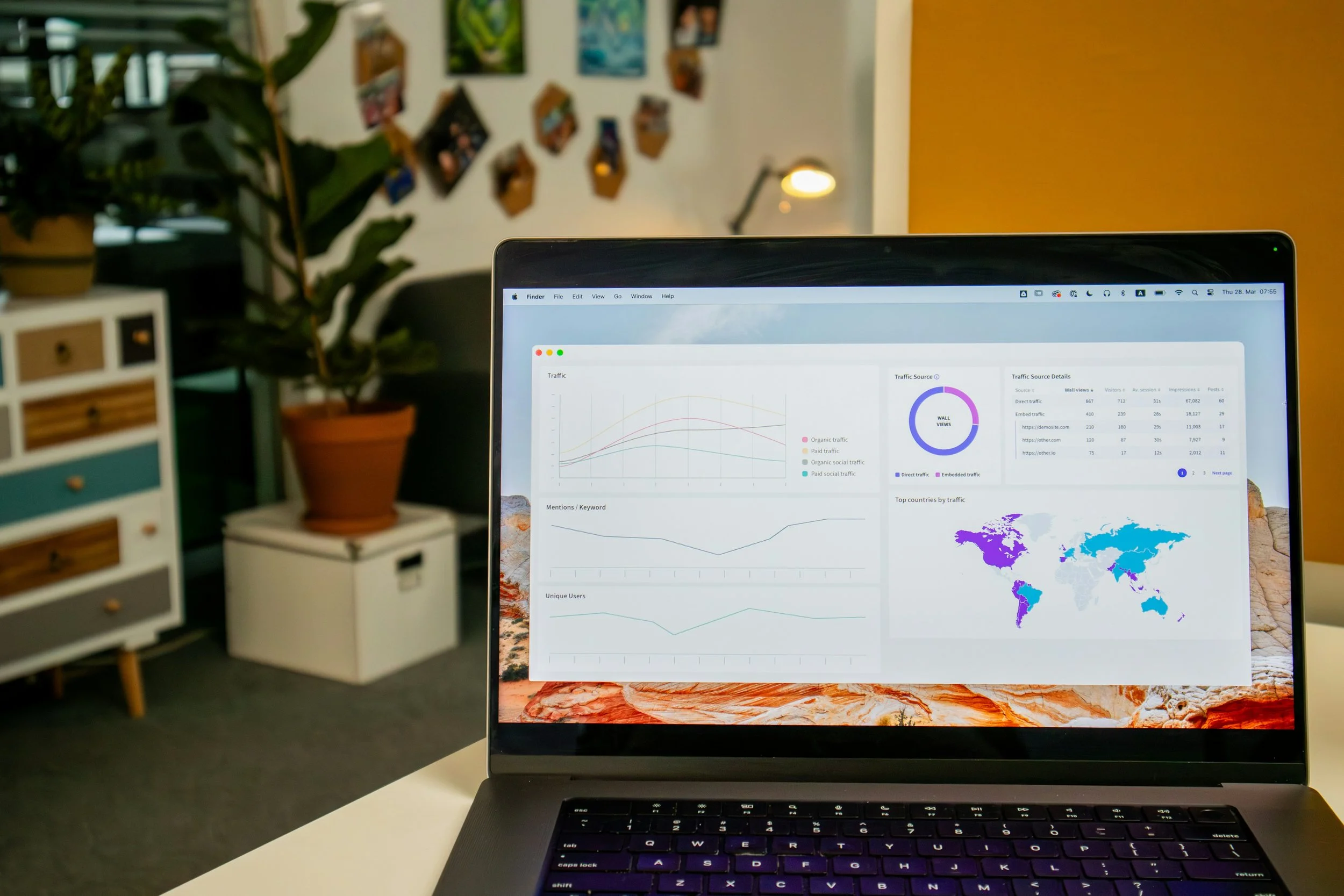 A laptop on a desk displaying analytics charts, world map, and traffic data, with a modern office space and plants in the background.