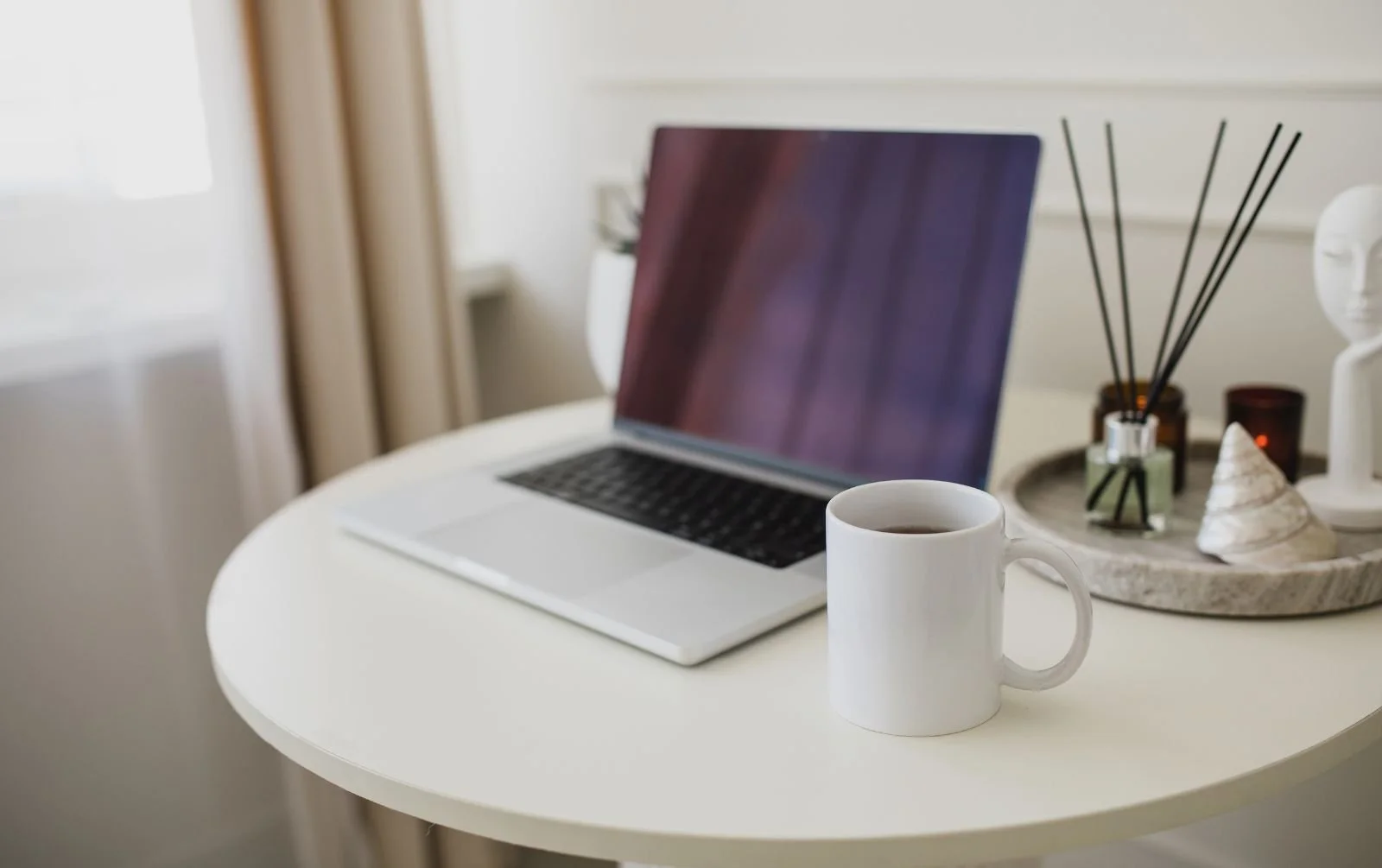 Open laptop and white mug on a round white table beside a decorative tray with a reed diffuser, candle and shell.
