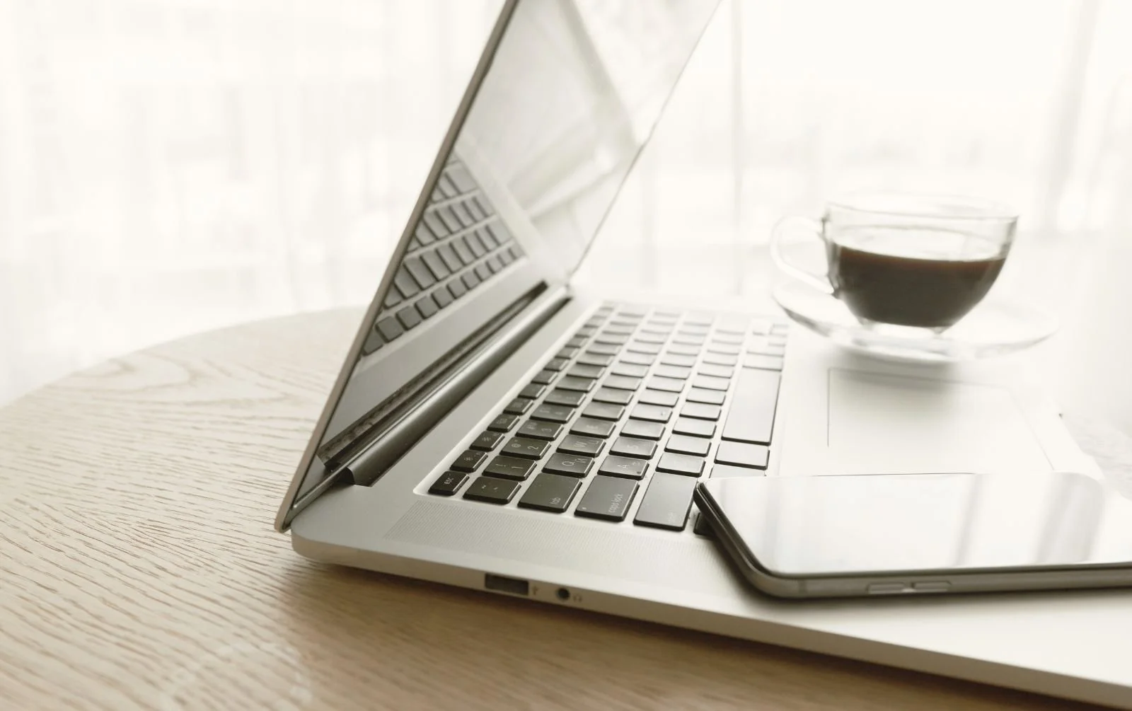 Open silver laptop with a smartphone and glass cup of black coffee on a wooden table beside a bright window.