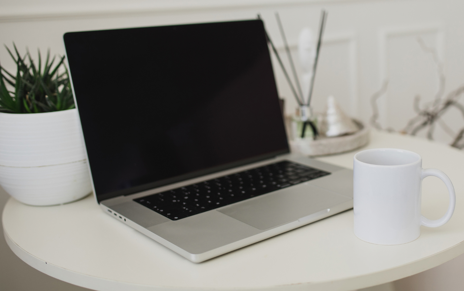 Laptop open on a white desk beside a plant, a reed diffuser and a white mug, ready for setting up a booking system and marketing automation