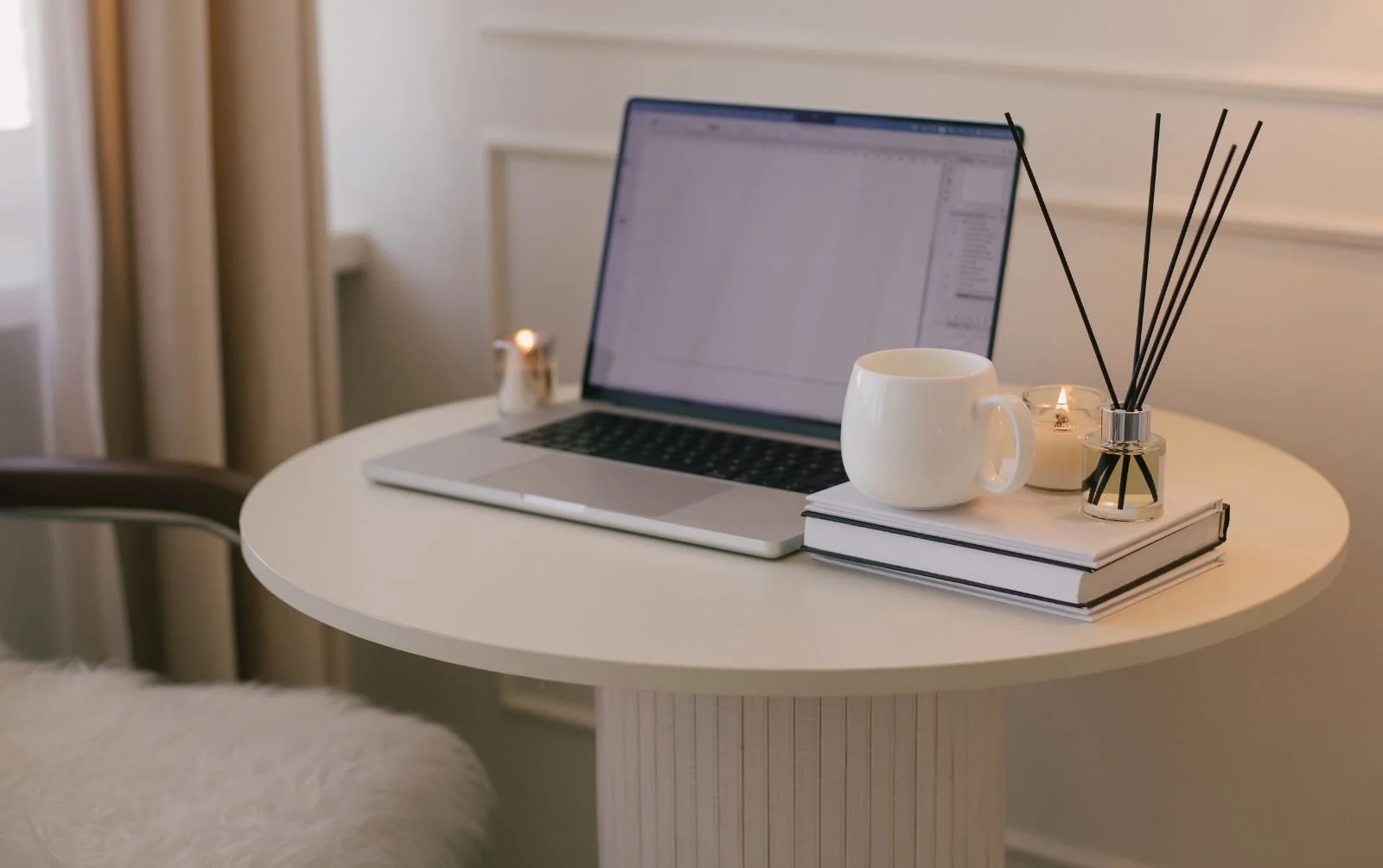 Laptop displaying design software on a round white table with stacked books, a white mug, lit candles and a reed diffuser.