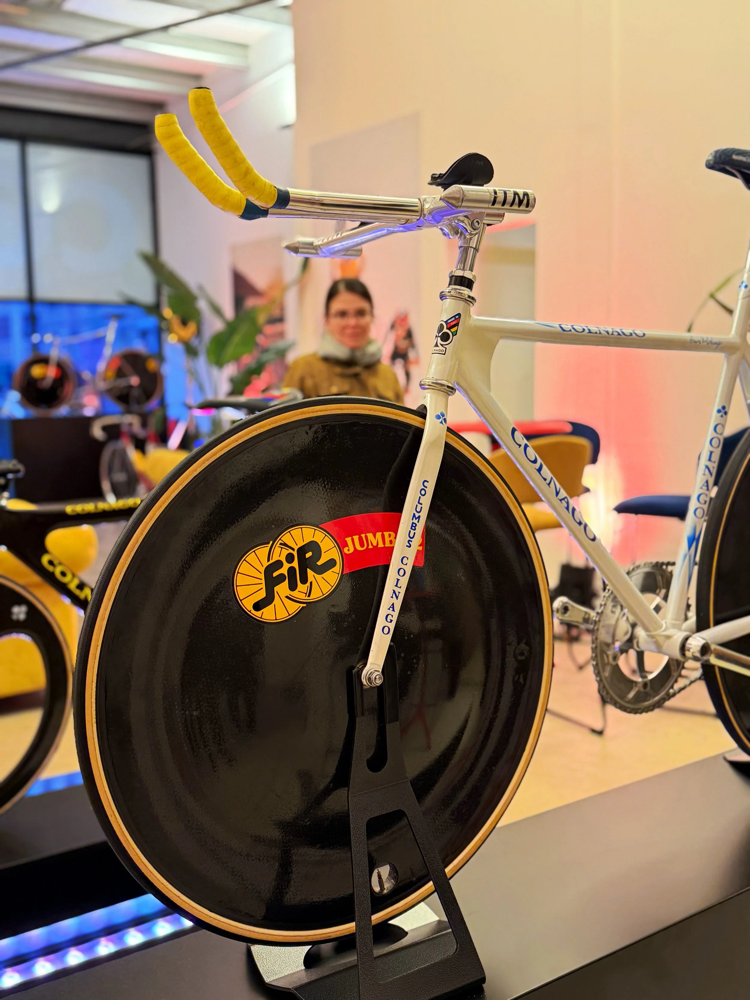 Close-up of a bicycle with a disc wheel, branded Colnago, on display. The bike has yellow handlebar tape and a white frame with blue accents. There is a woman in the background looking at the display in an indoor setting.