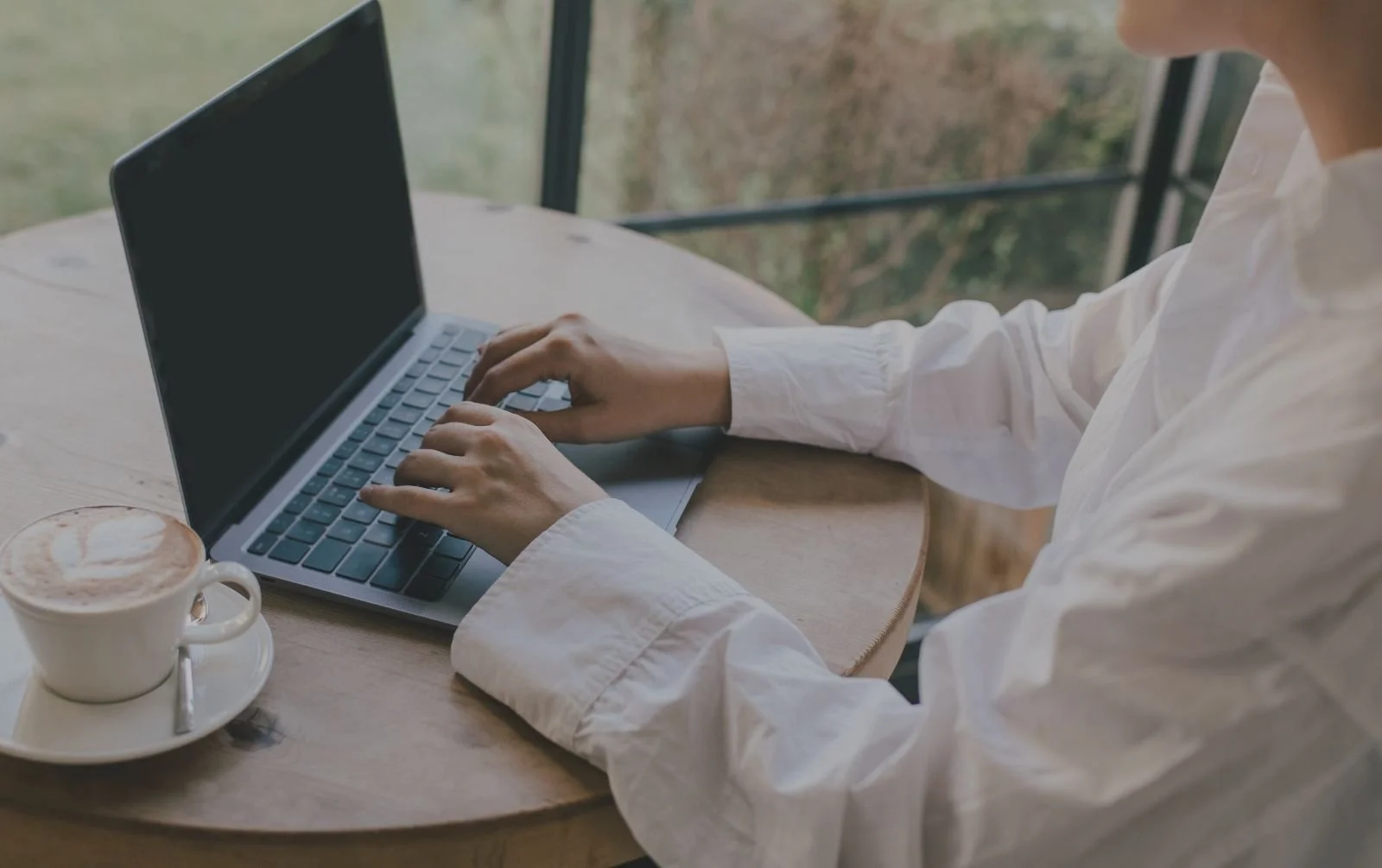 A person in a white shirt typing on a laptop at a round wooden table beside a window, with a cappuccino next to them.