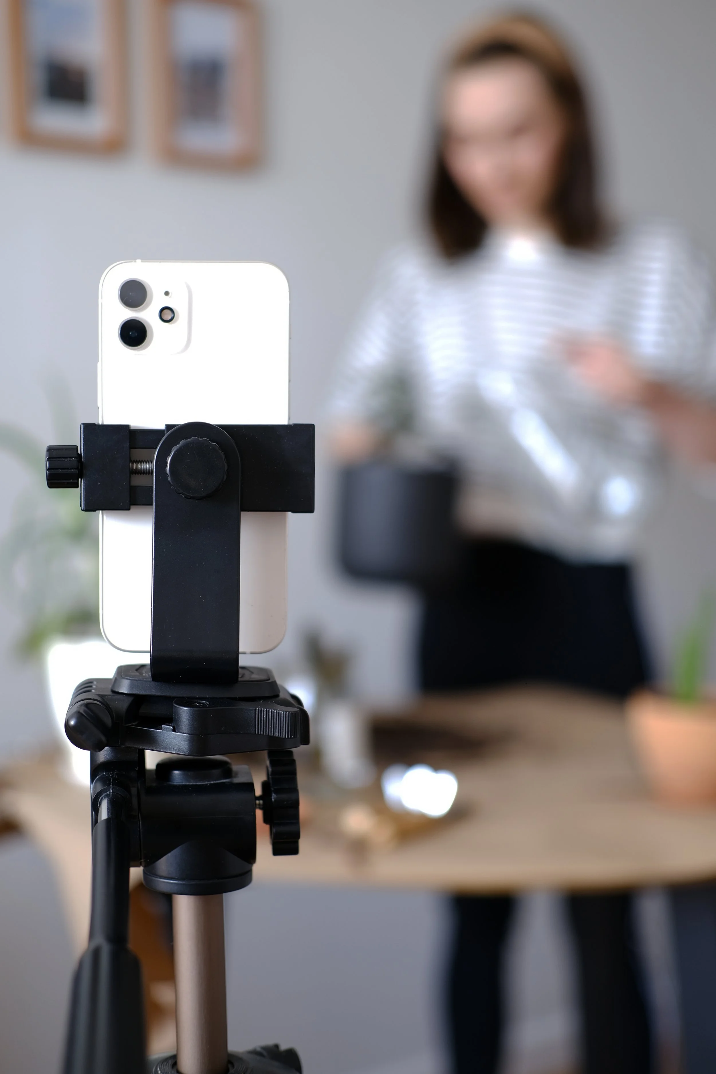 A smartphone mounted on a tripod, with a blurred woman in the background holding a cup and standing near a table with plants and decor items.