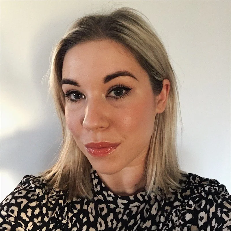A young woman with blonde hair and makeup, wearing a black and white patterned top, smiling slightly at the camera.