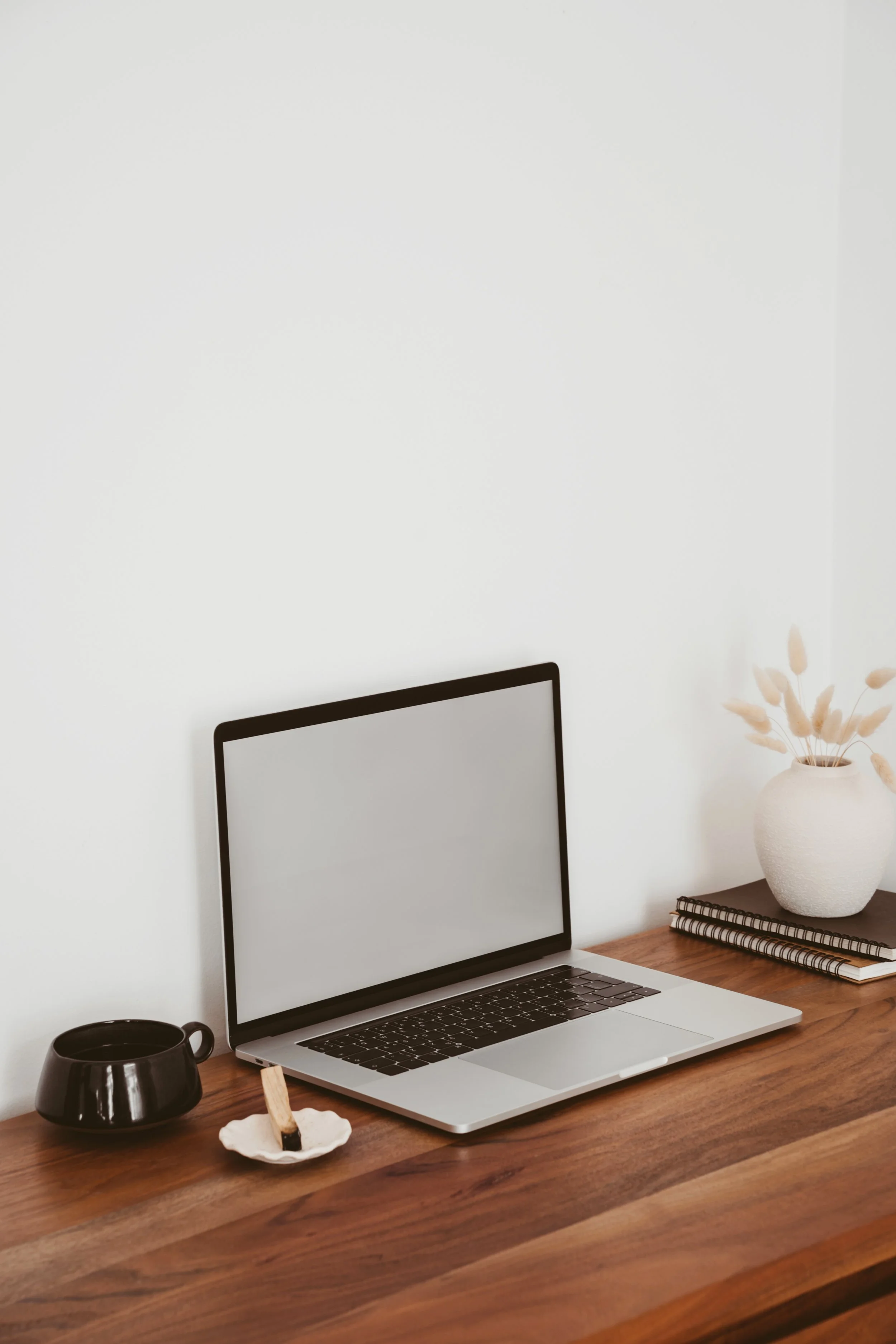 A minimalist desk setup with an open laptop, black mug, small white dish with a wood stick, a white vase with beige dried flowers, and a spiral notebook on a wooden table against a plain white wall.