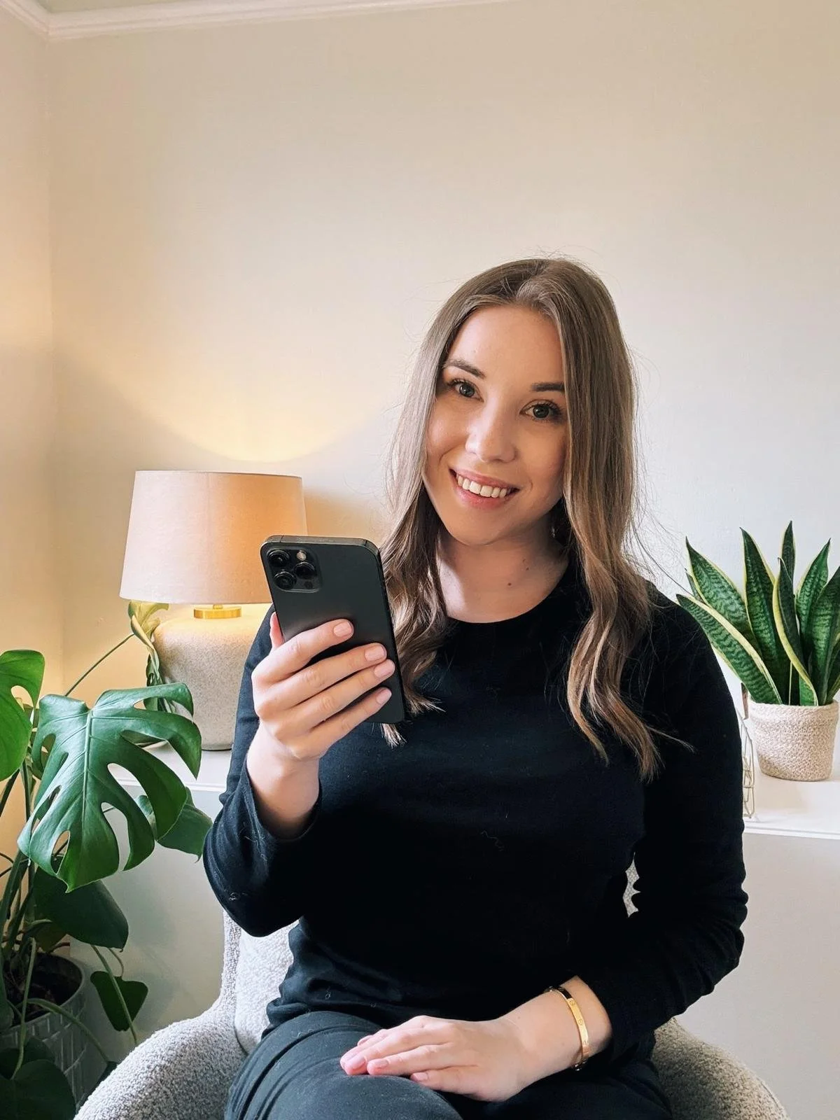 A young woman with long wavy brown hair and fair skin sitting on a beige armchair, smiling and holding a smartphone. She is wearing a black long-sleeve shirt, a gold bracelet on her wrist, and a black leggings. Behind her, there is a beige table lamp, a large green monstera plant, and a snake plant in a white pot, with a beige wall in the background.
