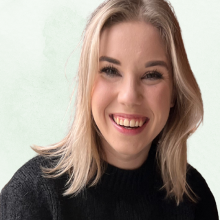 A young woman with blonde hair and a black sweater smiling in front of a light green background.