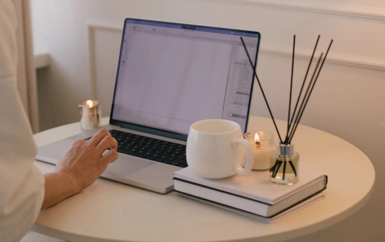 Person using design software on a laptop at a white table with a mug, stacked books, lit candles and a reed diffuser.
