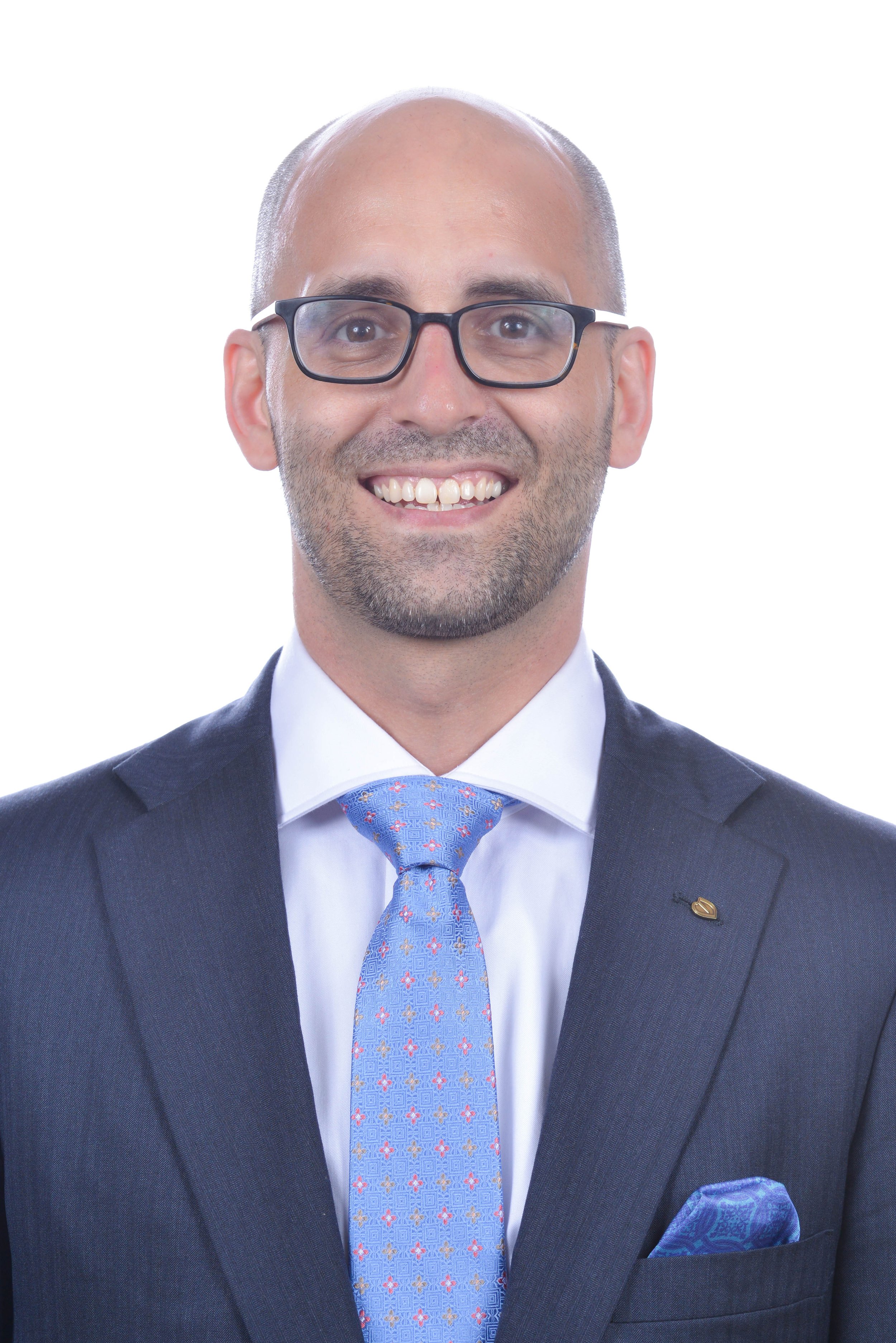 A smiling man wearing glasses, a dark suit, a white shirt, a light blue tie with red and yellow patterns, and a matching pocket square.