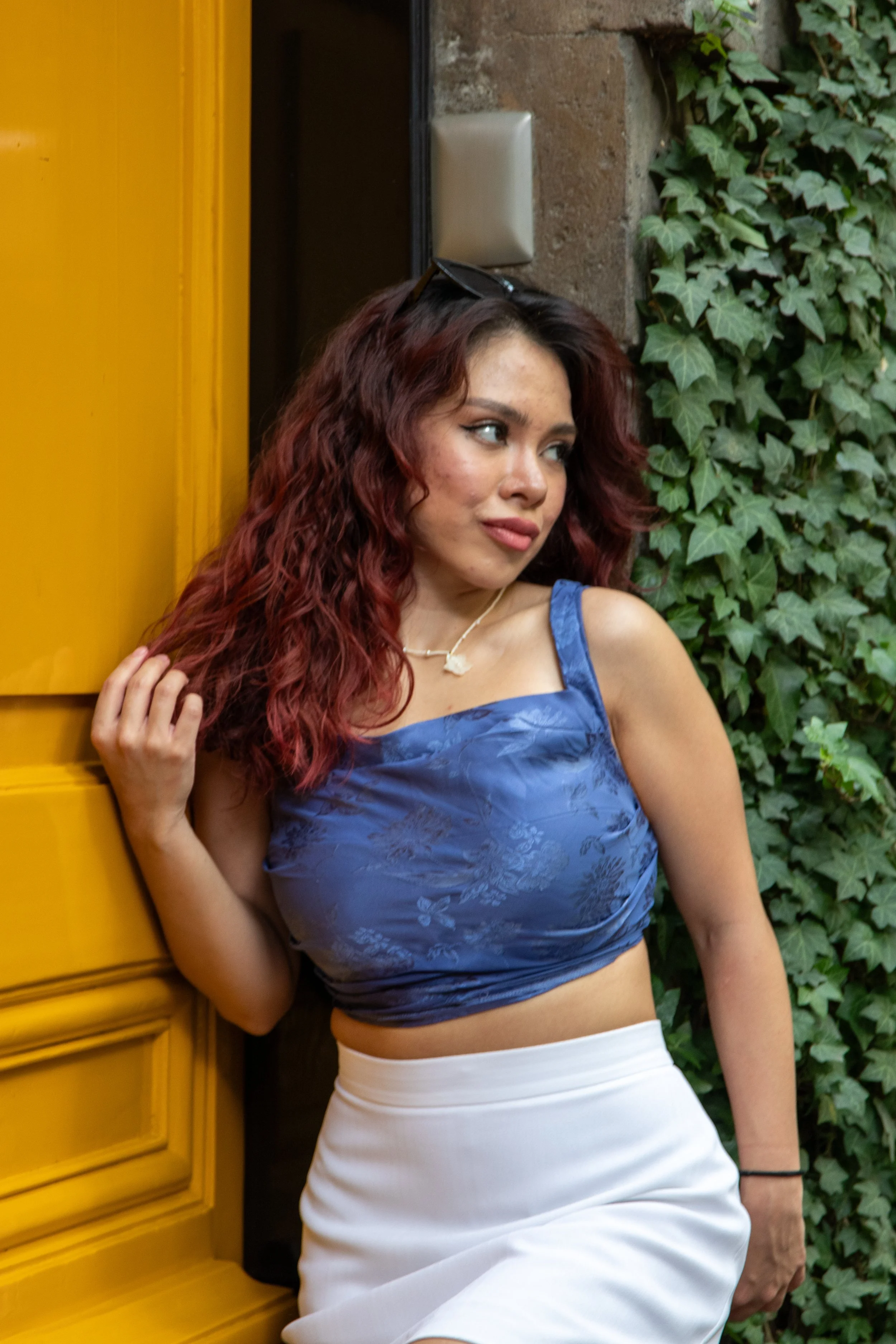 A woman with curly red hair wearing a blue crop top and white skirt, standing next to a yellow door and green ivy. She has sunglasses on her head and is touching her hair with one hand.