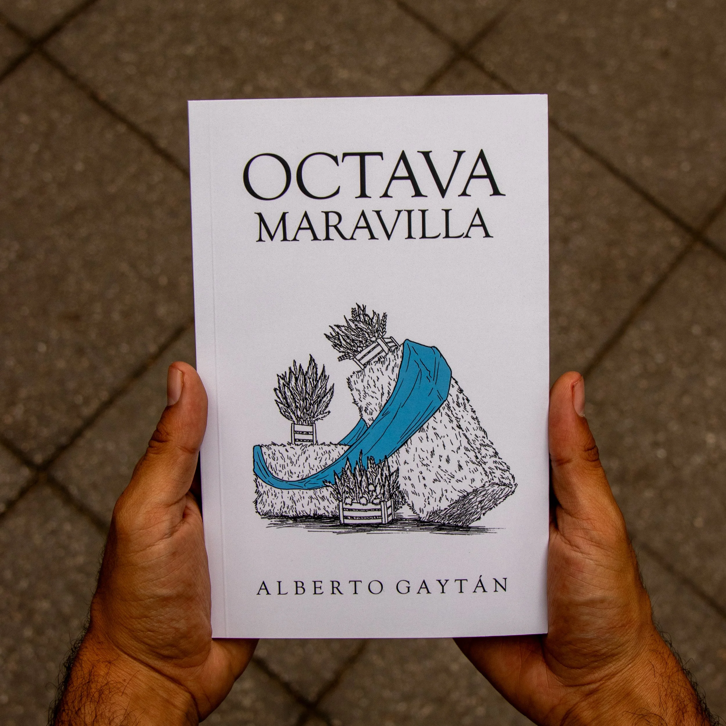 A person holding a book titled "Octava Maravilla" by Alberto Gaytán, with a line drawing of a character with plants and a mountain in the background, set against a tiled floor.