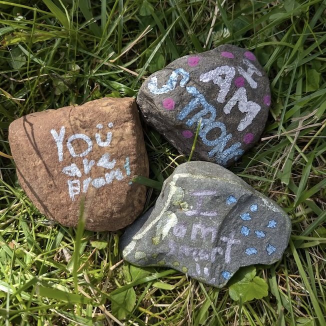 Three painted rocks with messages and designs, placed on green grass. One says "You are appreciated," the second has "Stay strong" with decorative elements, and the third says "I am important too" with colorful dots.