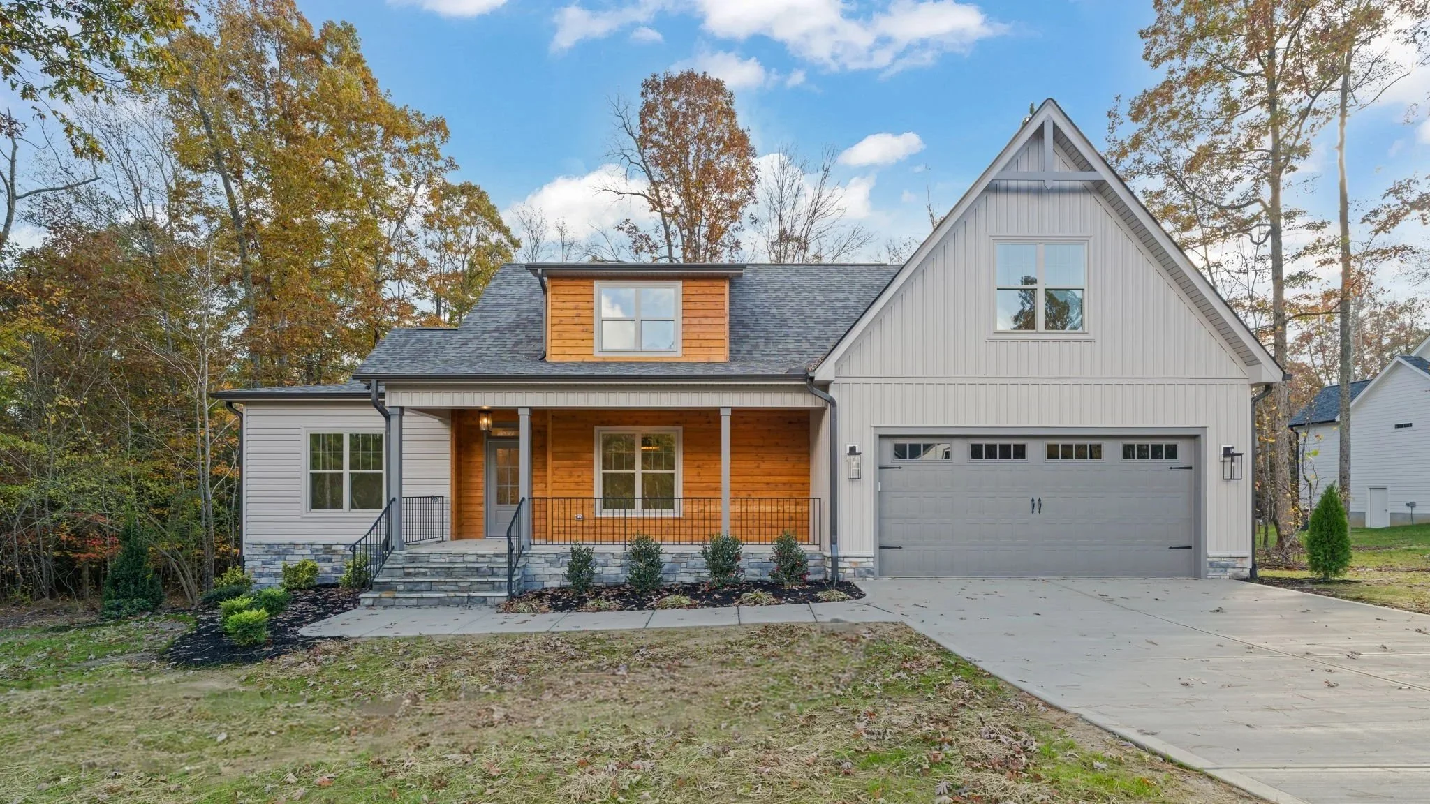 Front view of a modern two-story house with a gray garage door, wooden accents, a porch, steps, and some landscaped bushes, with trees in the background and a partly cloudy sky.