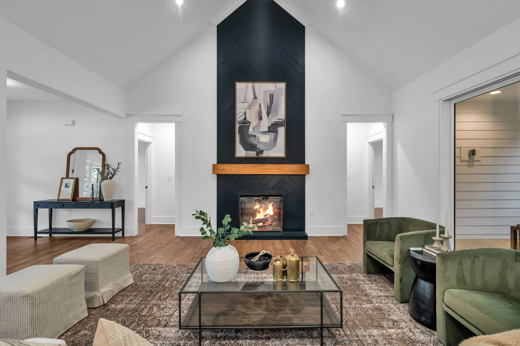 Living room with a high ceiling, a black fireplace with a wooden mantel, abstract artwork above, green armchairs, a glass coffee table with decorative items, and a side console with a mirror and vase.