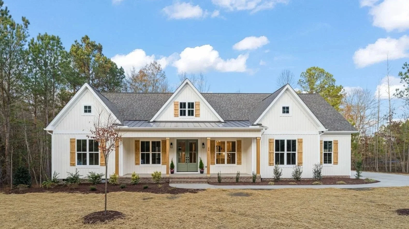 Modern farmhouse house with white vertical siding, wooden shutters, and a front porch, surrounded by a landscaped yard and trees under a blue sky.
