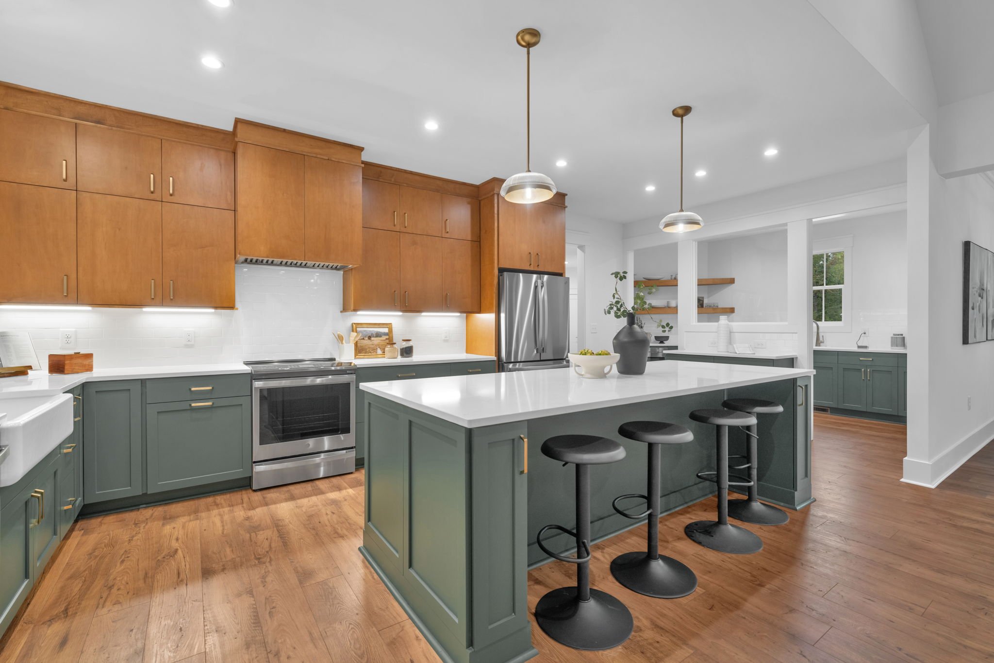 Modern kitchen with green cabinetry, wooden upper cabinets, white countertops, stainless steel appliances, and wooden flooring. There are three black barstools at the kitchen island, pendant lights, and open shelves with decorative items.