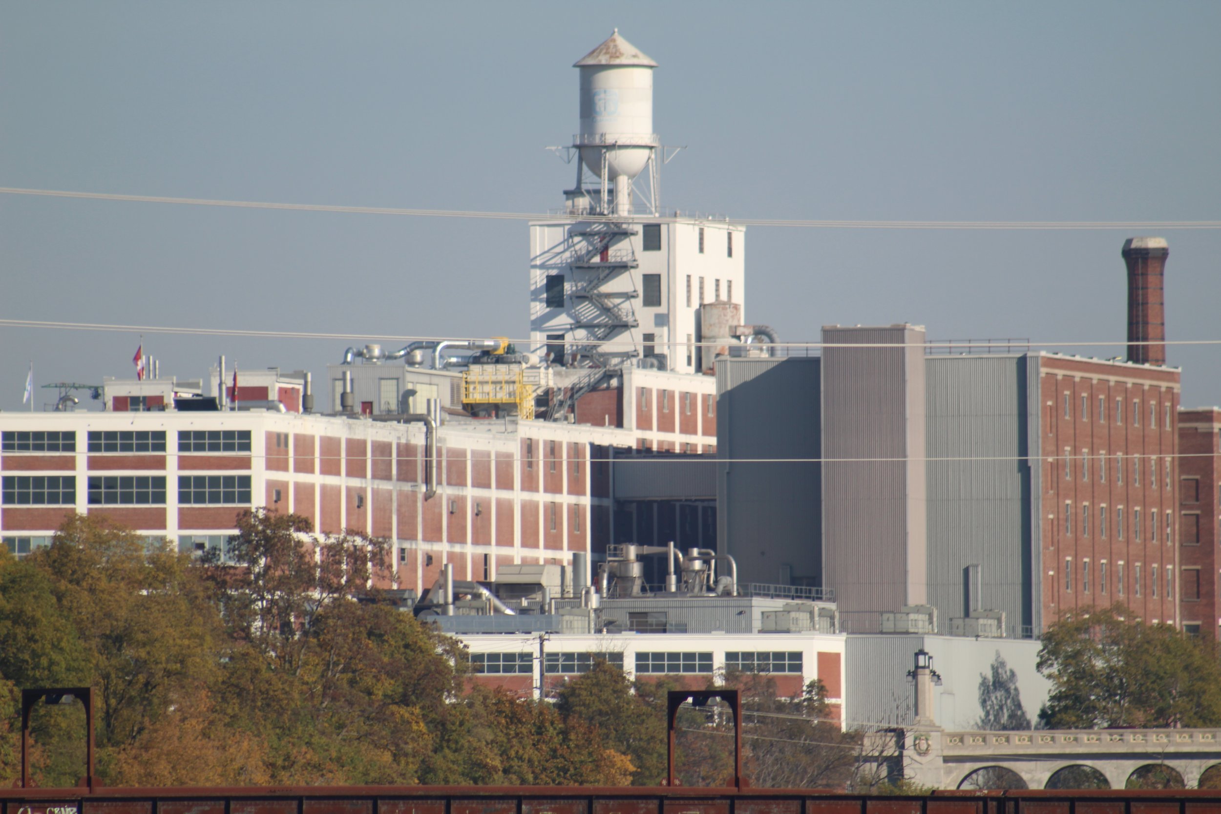 An industrial building with a white water tower on top, surrounded by brick and metal structures, with trees in the foreground and power lines crossing the scene.