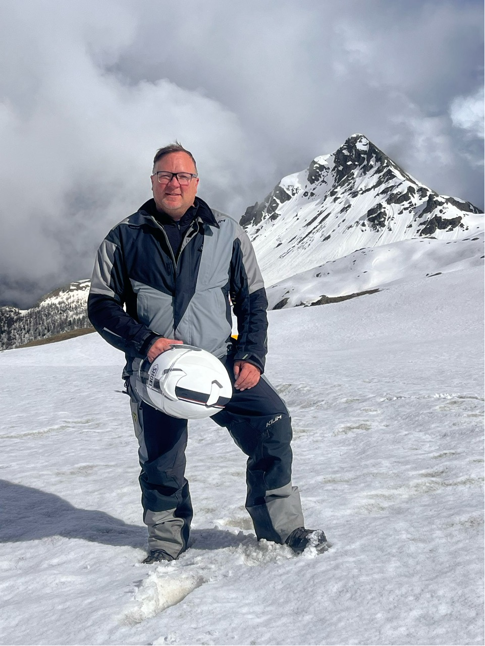 A man dressed in winter gear holding a white helmet, standing on snow with a mountain in the background.