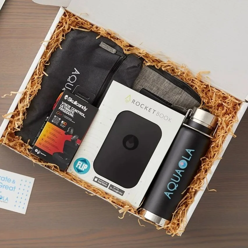 Gift box containing an Aqualan insulated water bottle, Skullcandy wireless earbuds, a Rocketbook smart notebook, and a black carrying case, all arranged on shredded paper in a gift box.