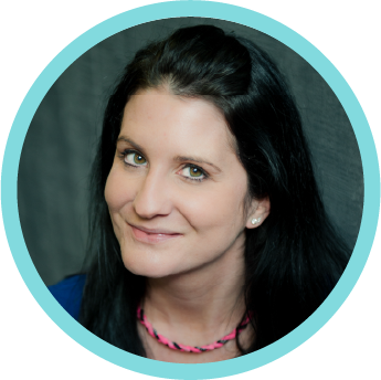 A woman with dark hair smiling at the camera, wearing a blue top and a pink necklace.