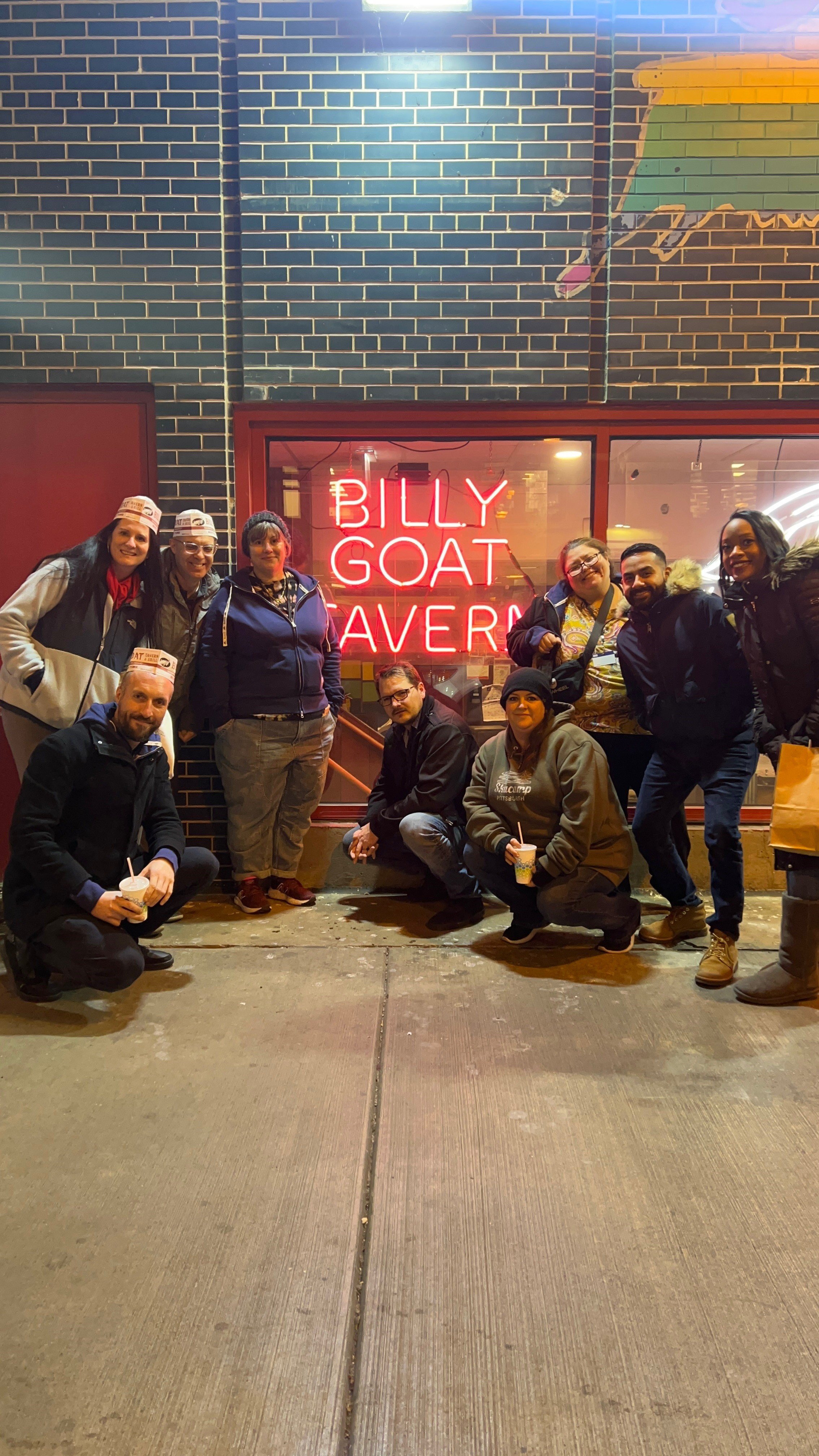 A group of nine people posing outside a restaurant at night, in front of a neon sign that reads 'Billy Goat Tavern'. They are dressed in casual winter clothing and some are holding ice cream cones.