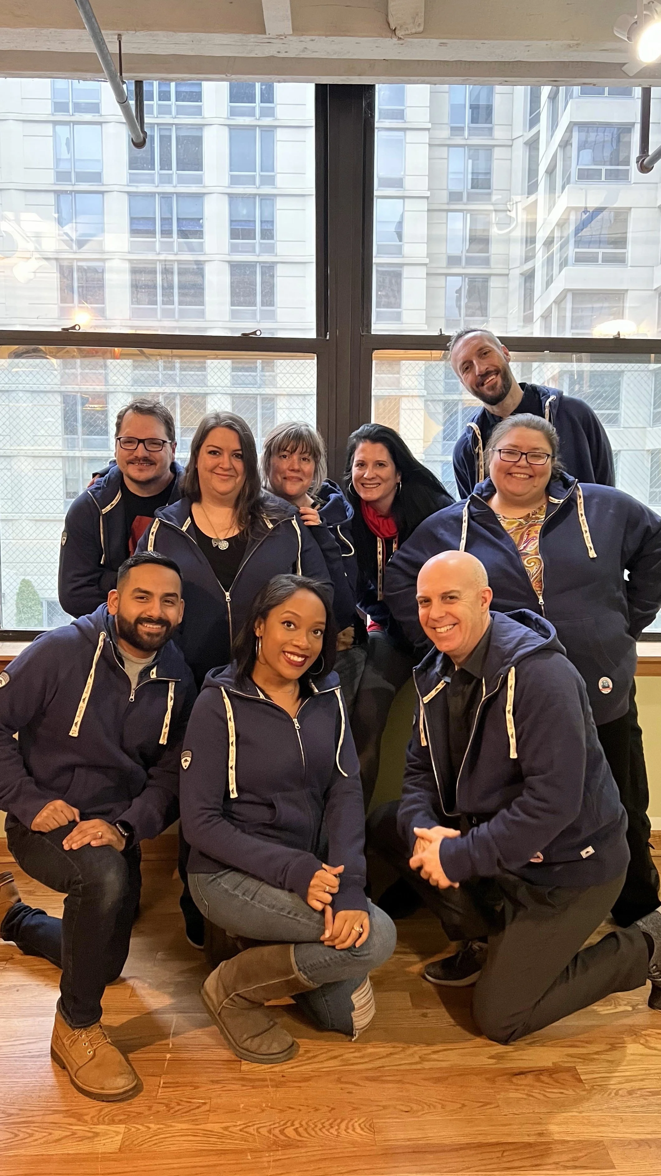 A group of nine people posing together indoors in front of large windows with a cityscape background. Everyone is wearing matching navy blue jackets with yellow accents, smiling at the camera.