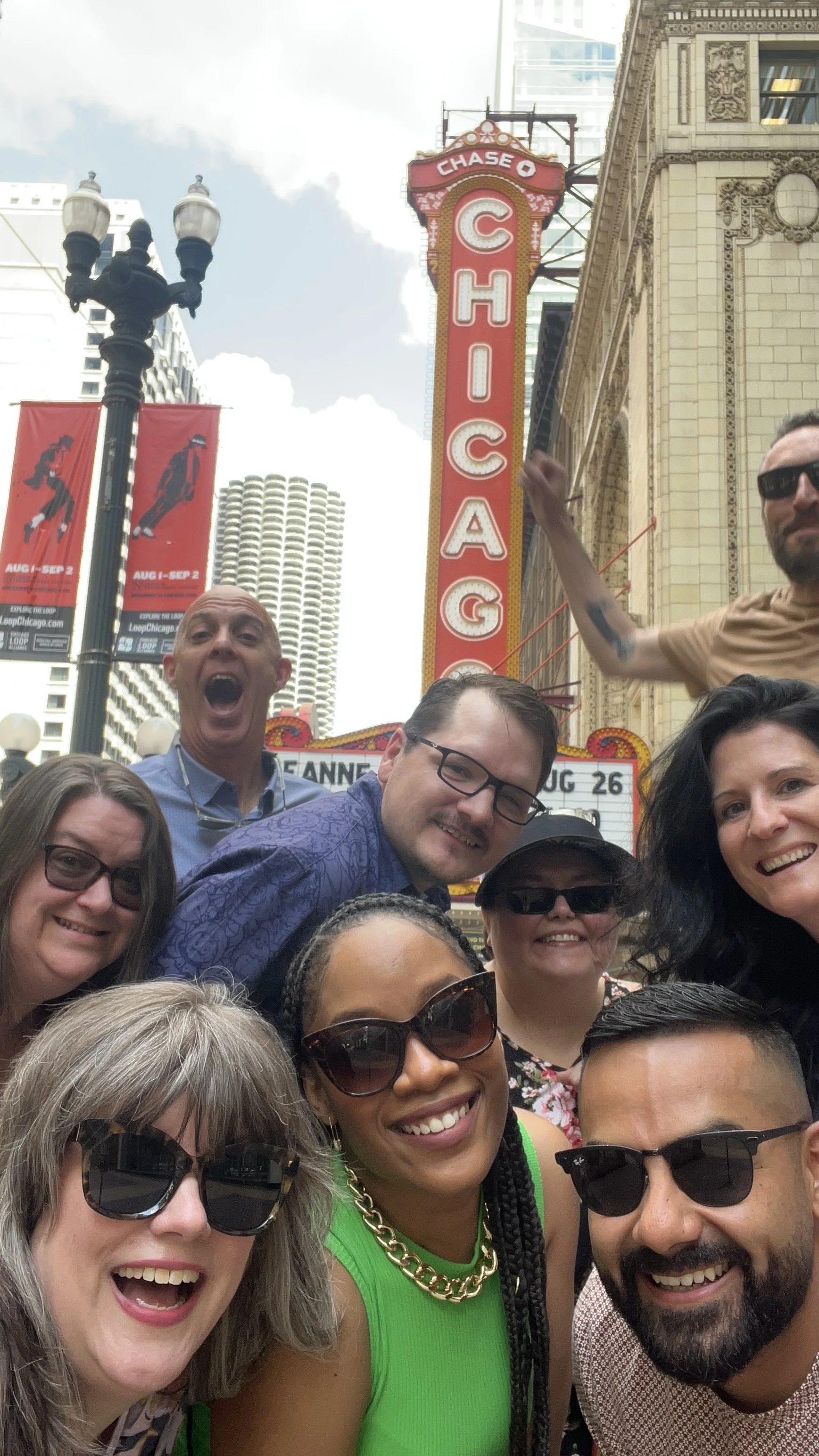 A group of people taking a selfie in front of the Chicago Theatre marquee. The marquee reads 'Chicago' and features a vintage design.