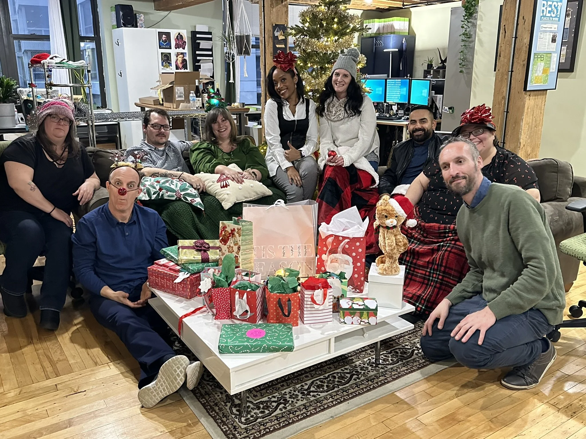 Group of people celebrating Christmas in an office, sitting and kneeling around a table with wrapped gifts, a teddy bear, and holiday decorations. Some wear holiday hats and sweaters, with a Christmas tree in the background.