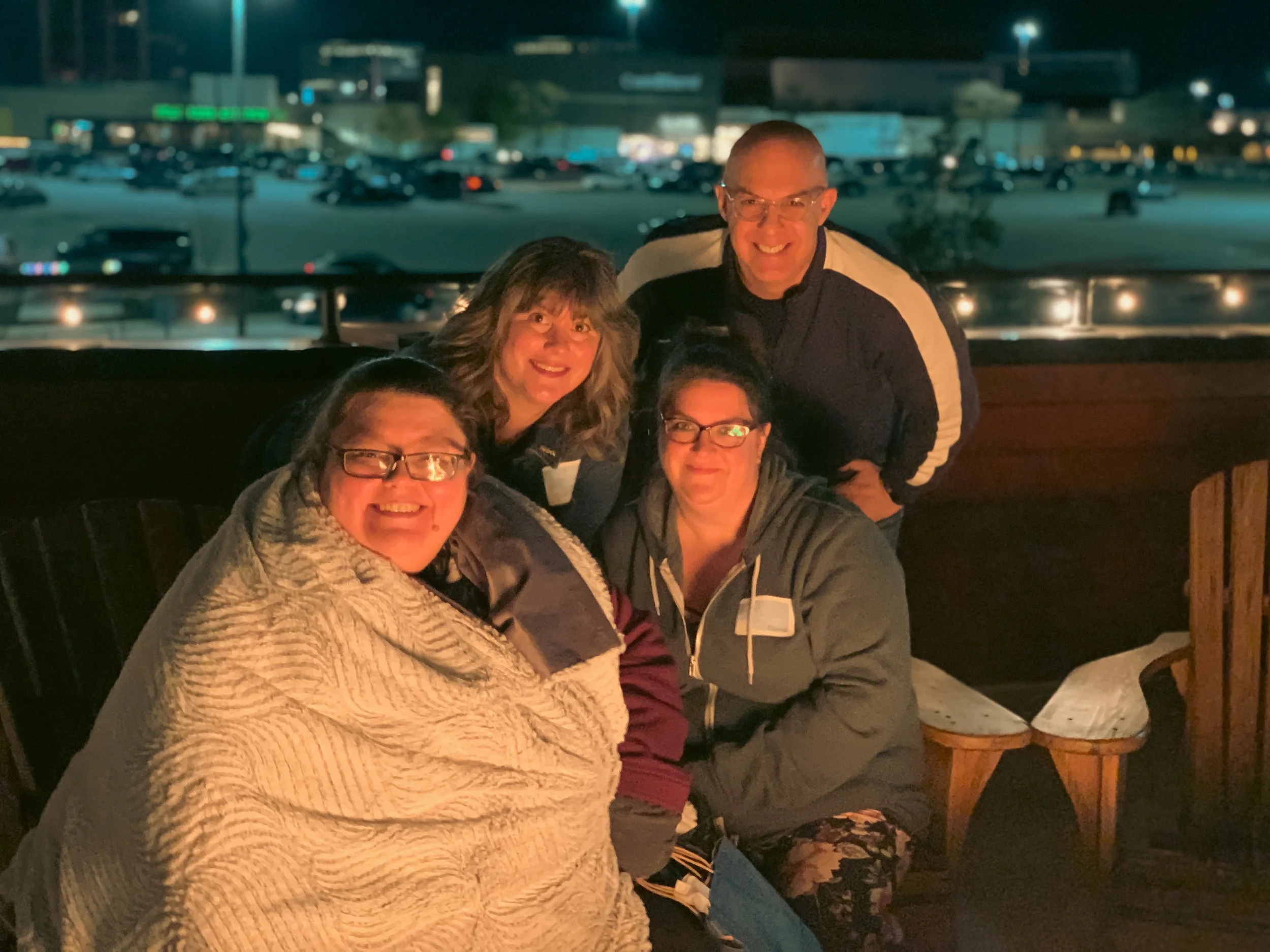 Four people sitting and standing together in a dimly lit indoor setting, possibly a restaurant or bar, with a window showing a parking lot and buildings outside at night.
