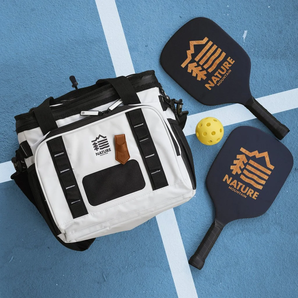 A white and black sports bag, two pickleball paddles with the word "NATURE" and mountain logo, and a yellow pickleball ball on a blue pickleball court.