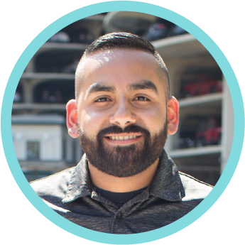 A close-up of a man with short dark hair and a beard, smiling, wearing a dark shirt, with a parking lot in the background.
