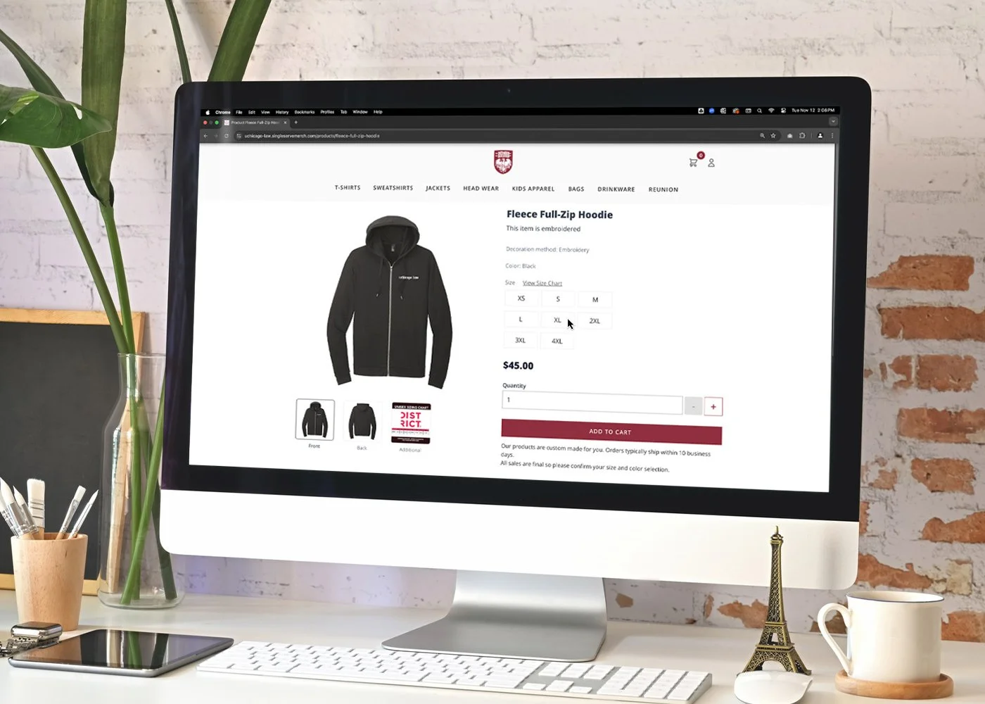 An iMac on a desk displaying an online shopping webpage for a black fleece full-zip hoodie. The screen shows hoodie details, size options, and price. Next to the iMac are a plant, a mug, a tablet, and a small Eiffel Tower figurine on the desk.