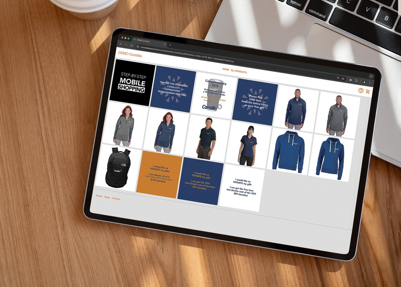 Tablet displaying an online store with apparel and merchandise, including jackets, backpacks, and promotional messages, on a wooden desk with a laptop in the background.